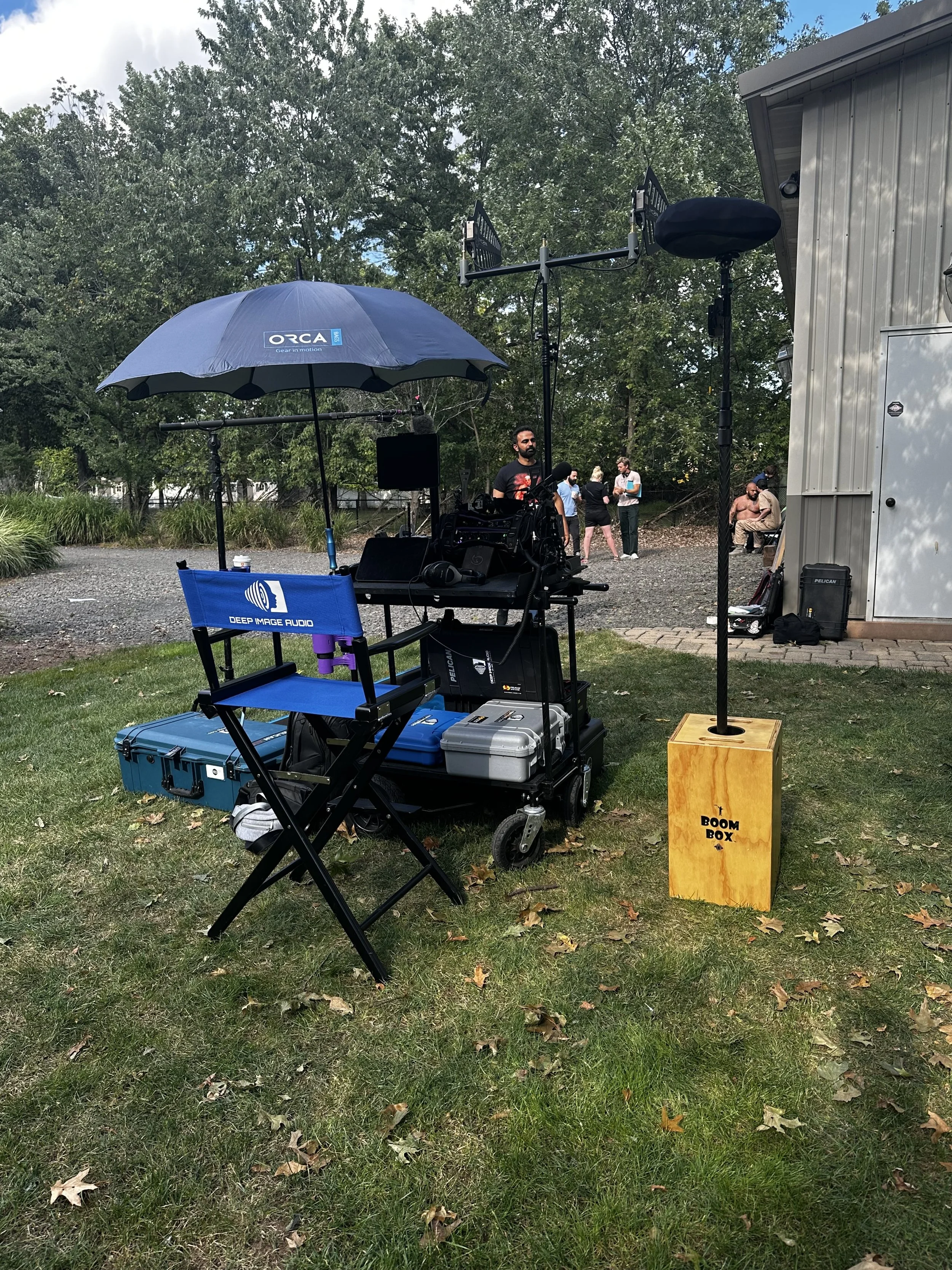 A videography or photography setup outdoors featuring a large umbrella with a logo, a chair labeled 'Deep Image Audio', equipment cases, and a small yellow box labeled 'Boom Box'. In the background, people are gathered near a building and trees.