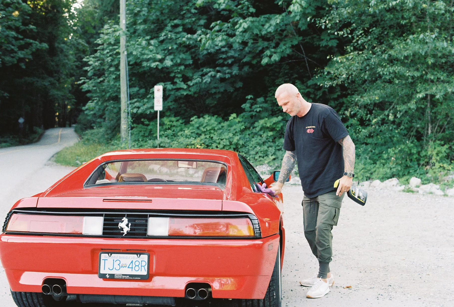 Photographer standing beside the rear of a red Ferrari 348 on a forest road, captured on Nikomat film.