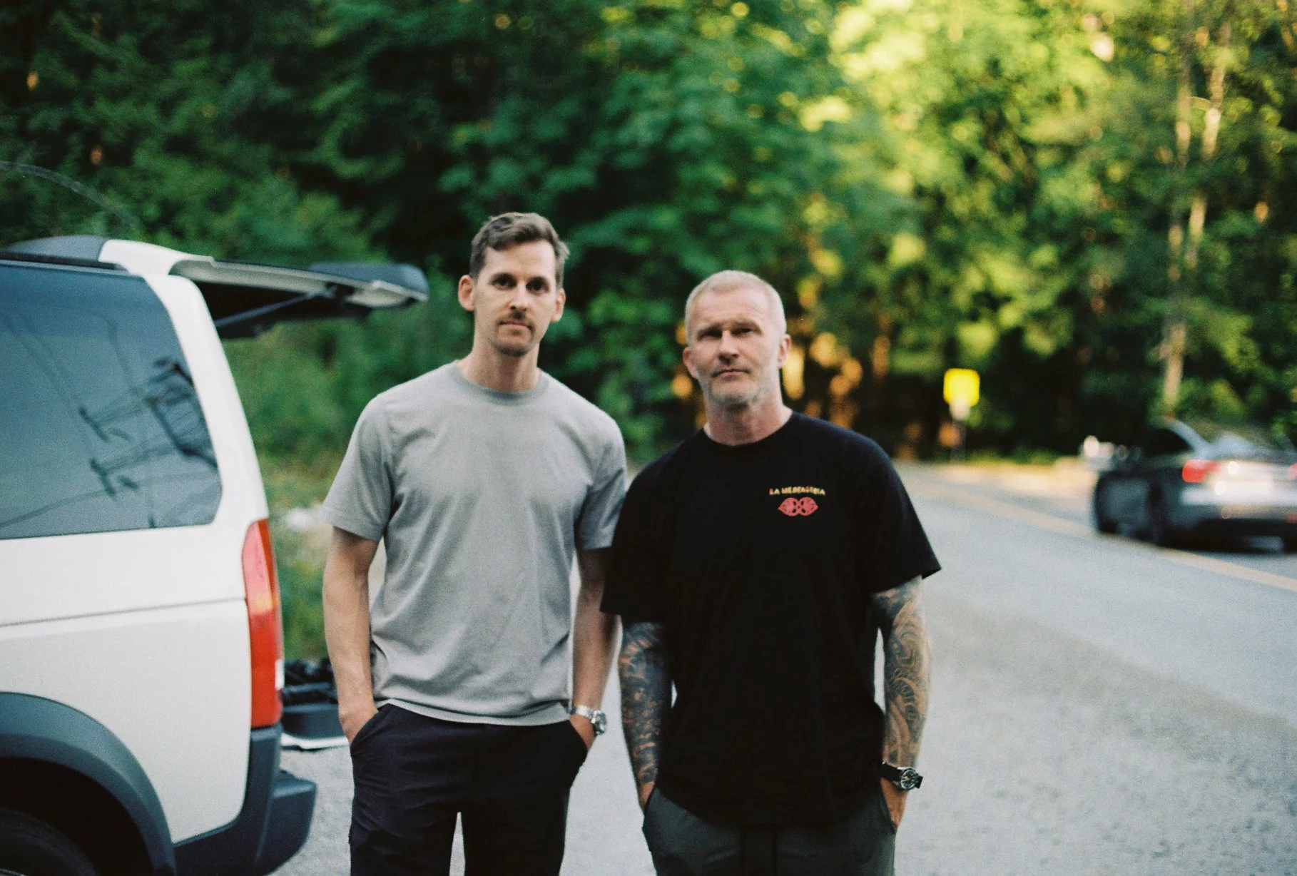 Two men standing beside a white vehicle on a tree-lined road, photographed on Kodacolor 200 with a Nikomat.