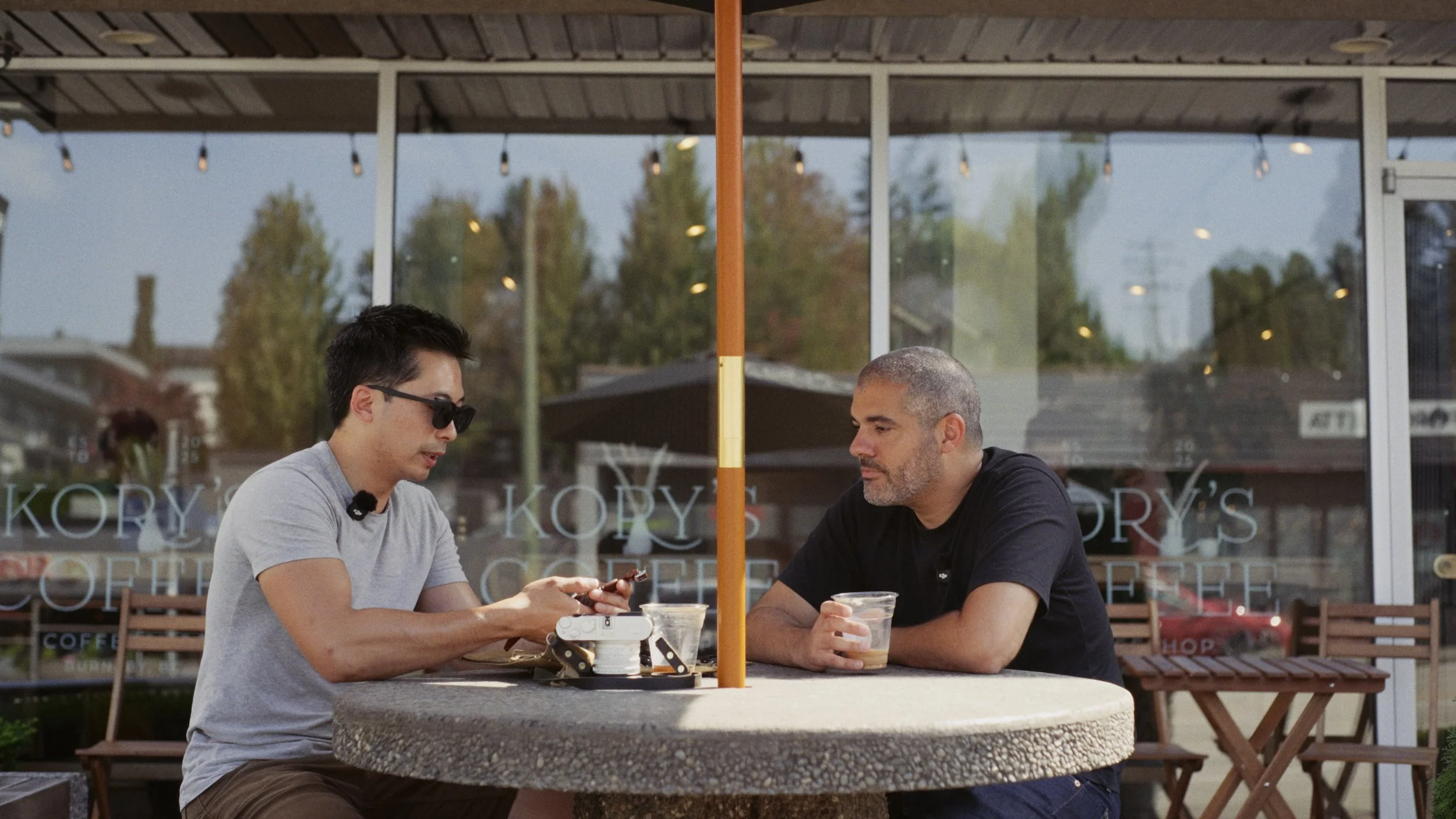 Julian Fok and Nima discussing a watch over coffee at an outdoor cafe table in a frame from the film.