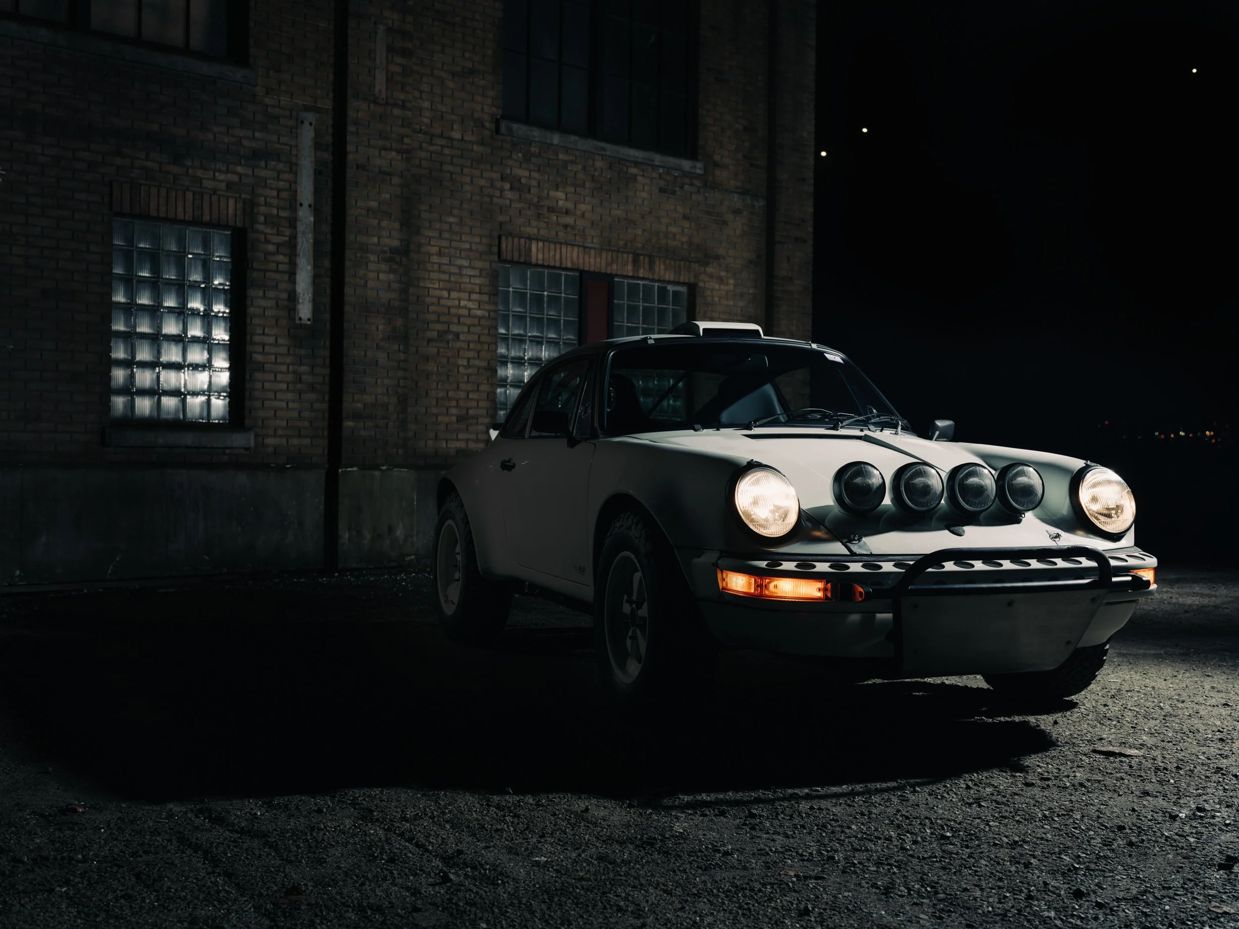 Three-quarter front view of Safari Porsche 911 with rally lights illuminated, captured near an old building at night.