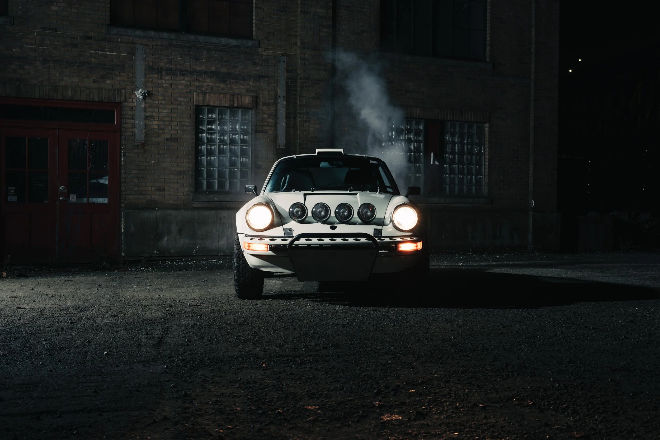 Front view of Safari Porsche 911 by Flatsix Classics. Headlights on, smoke rising from the engine, shot in an industrial area at night. Captured on Fujifilm GFX 100 ii