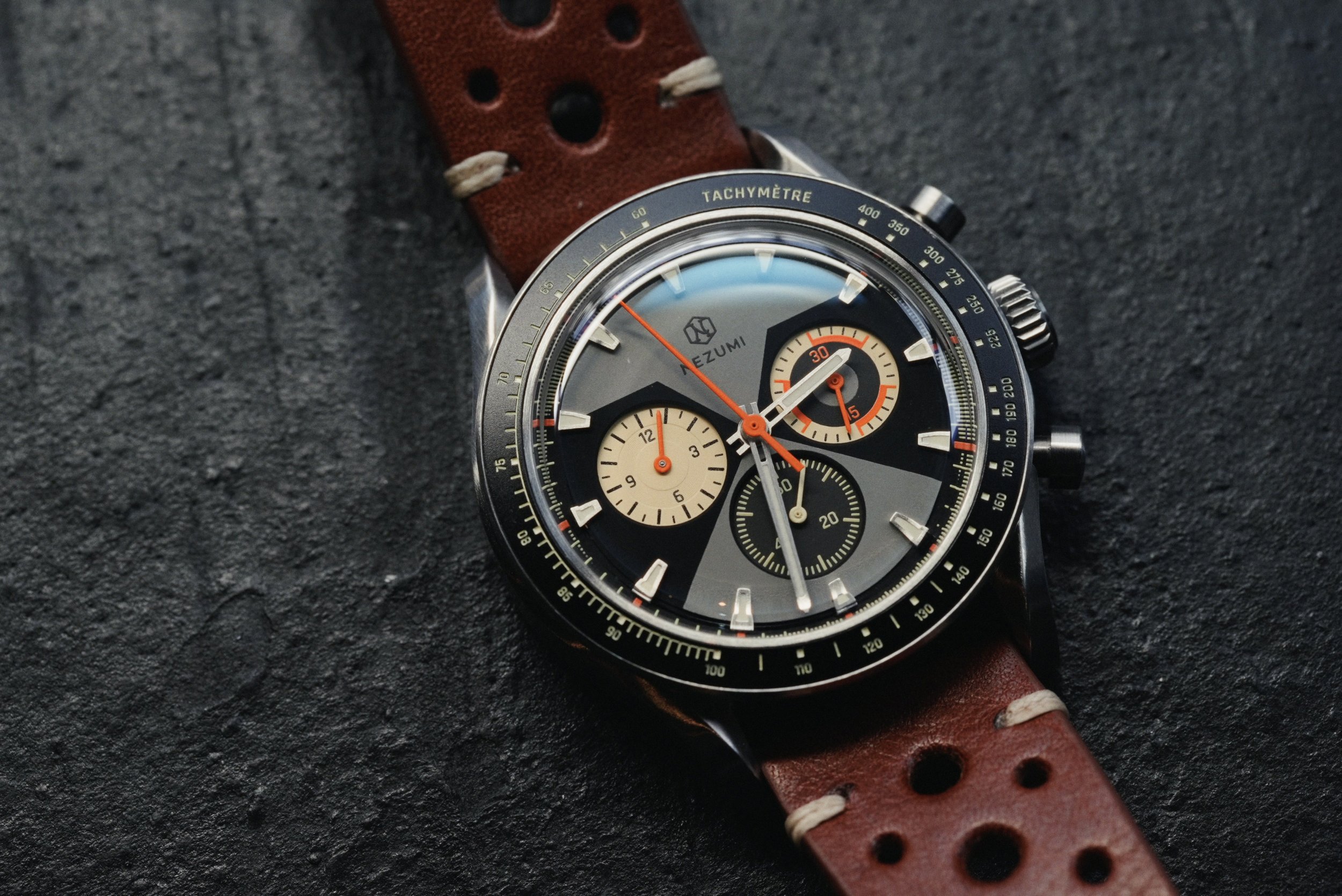 Close-up of the Nezumi Voiture VM2 dial, orange chronograph hands and perforated brown leather strap on a dark surface.