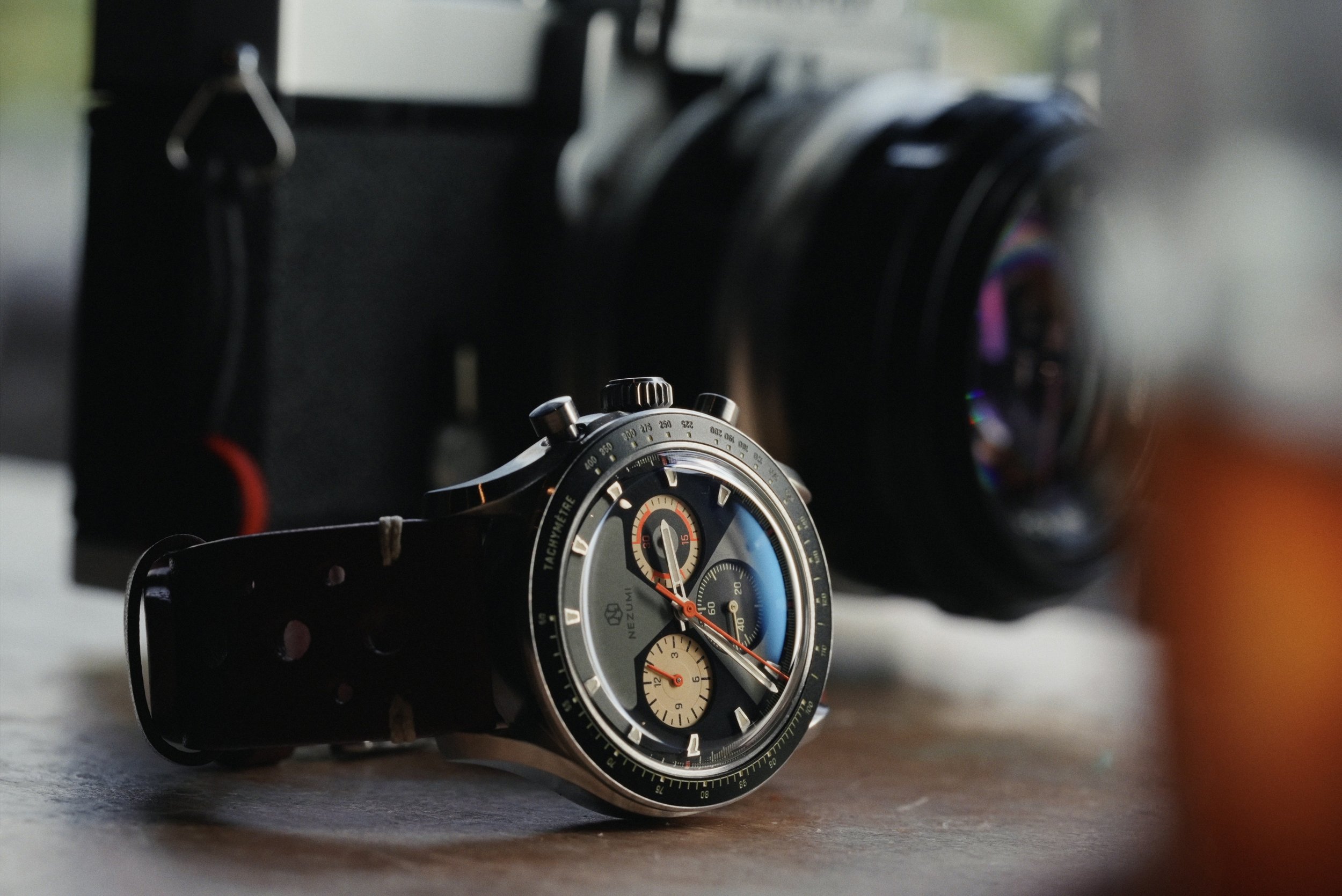 Nezumi Voiture VM2 chronograph resting on a table in front of a Nikomat film camera and lens.