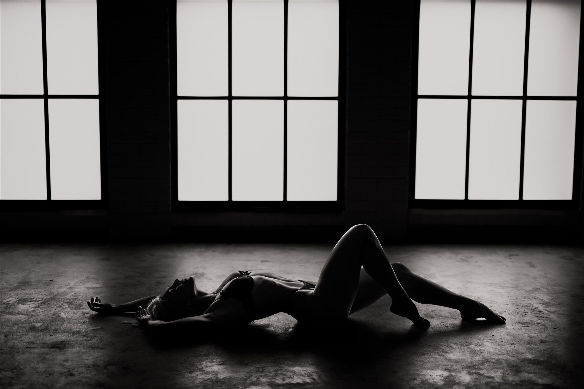 Silhouette of a woman lying on her back with one leg bent, in a dimly lit room with large windows in the background.