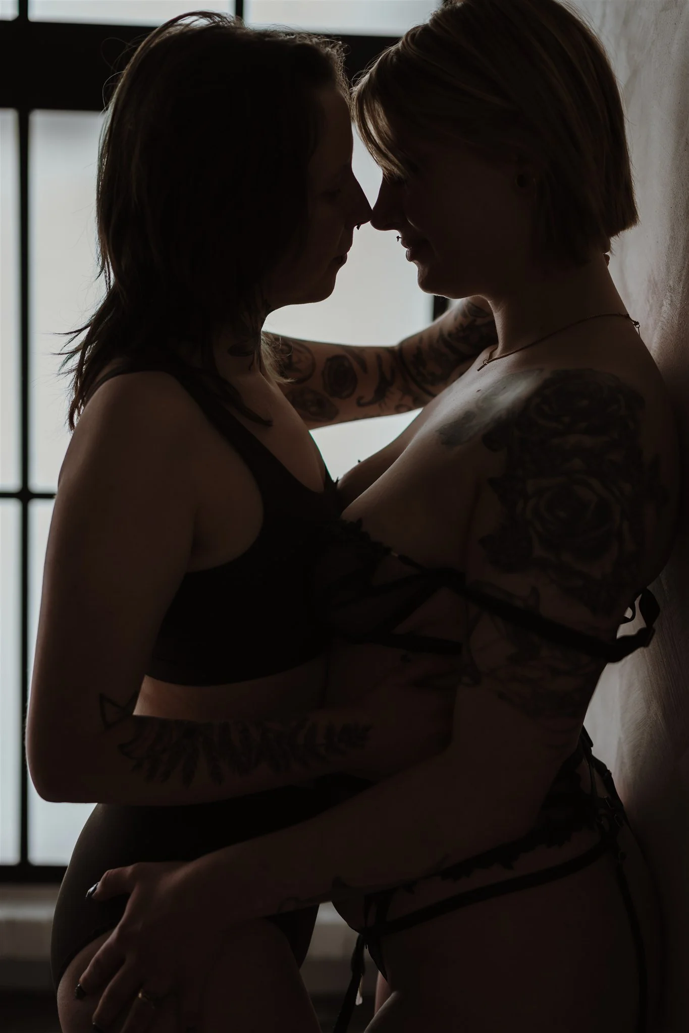 Two women with tattoos are close together, touching foreheads and smiling, in front of a window with grid patterns, in silhouette.
