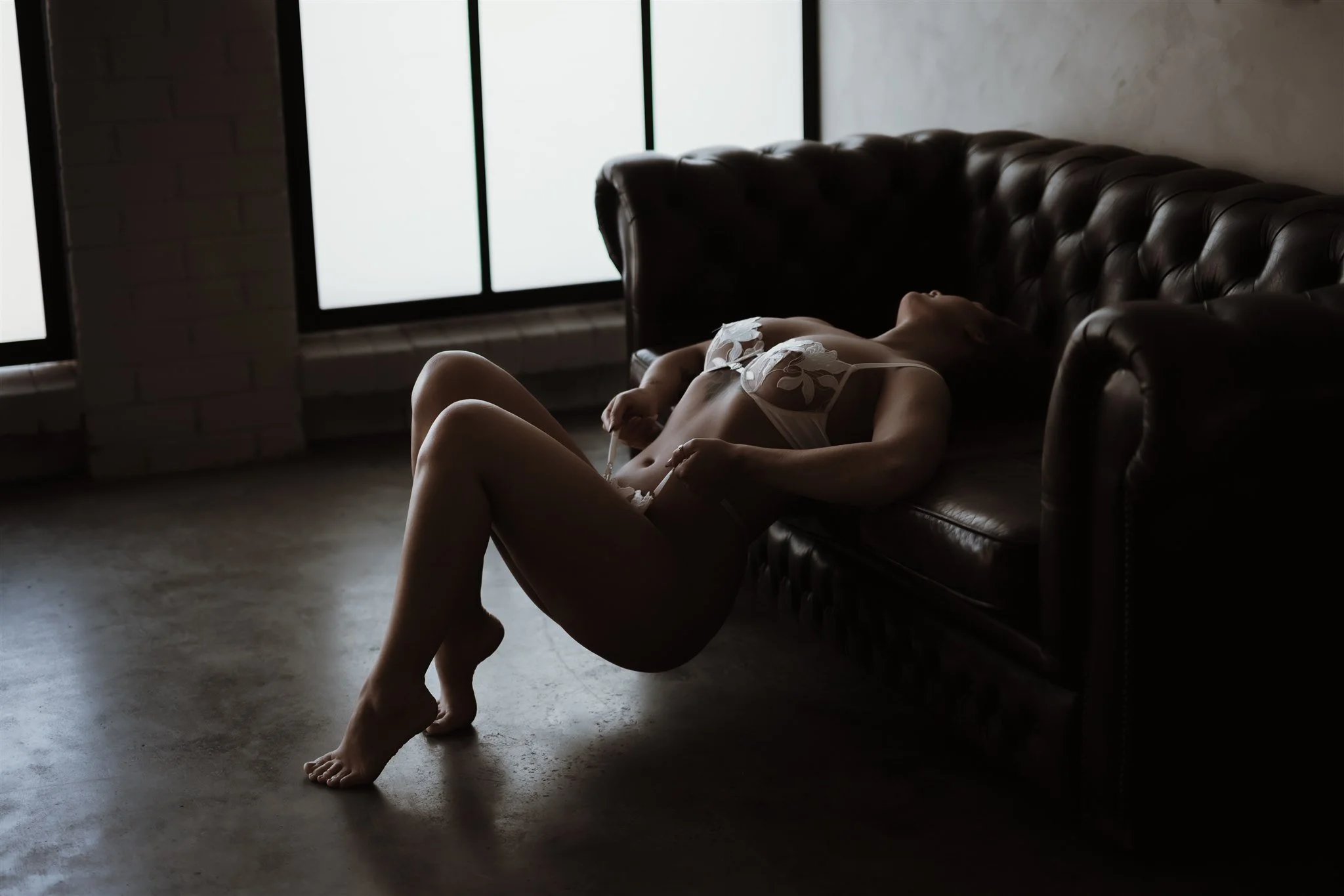 A woman in lingerie reclining on a dark leather couch in front of large windows with frosted glass, in a dimly lit room.