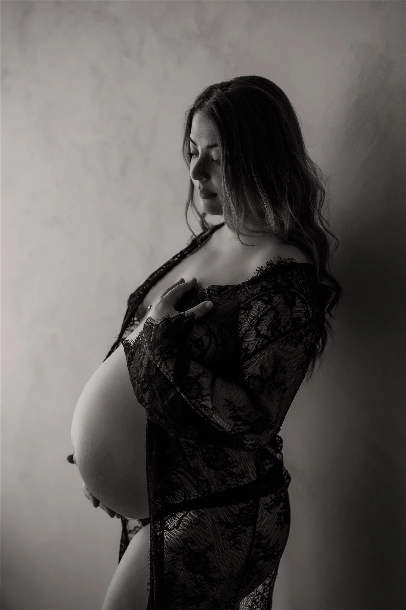 A pregnant woman with long wavy hair wearing a black lace robe, gently touching her belly, looking down with her eyes closed, against a plain background, black and white photo.