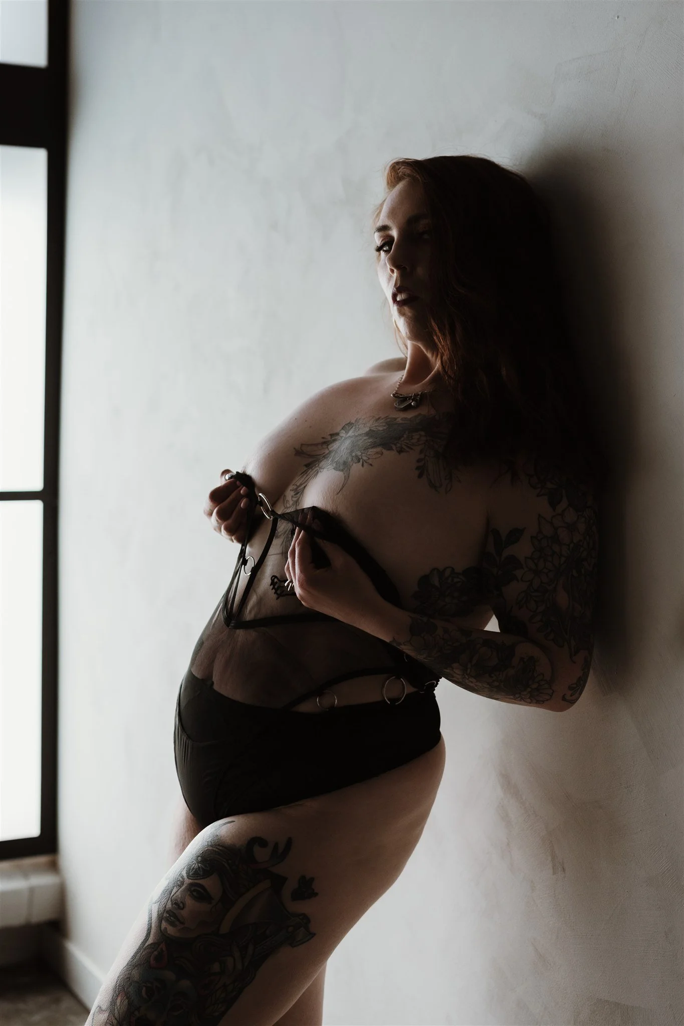 A woman with tattoos on her arm and thigh, wearing black lingerie, standing against a plain wall near a window, looking thoughtfully to the side.