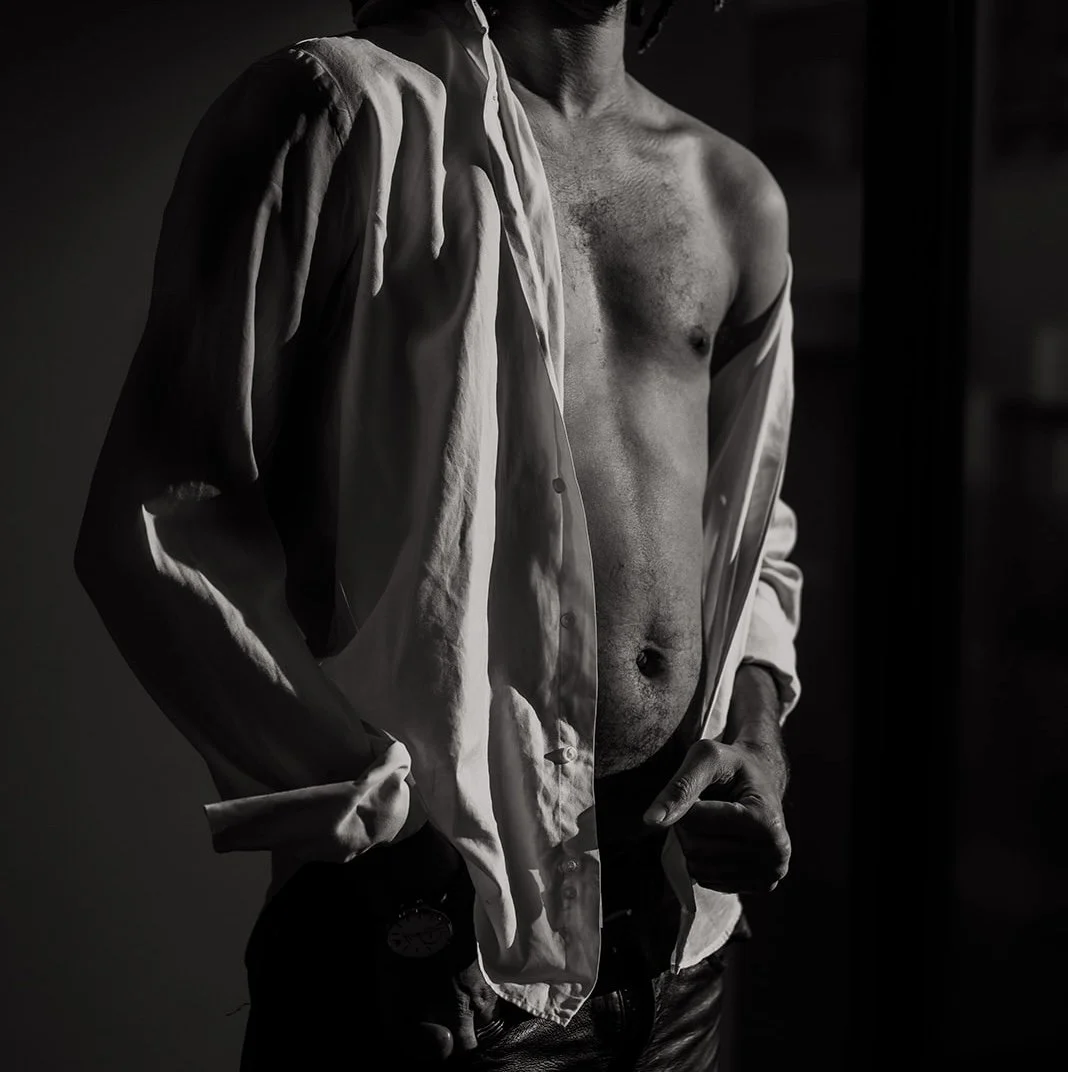 A man with an open shirt draped over his shoulder, standing in a dimly lit room.