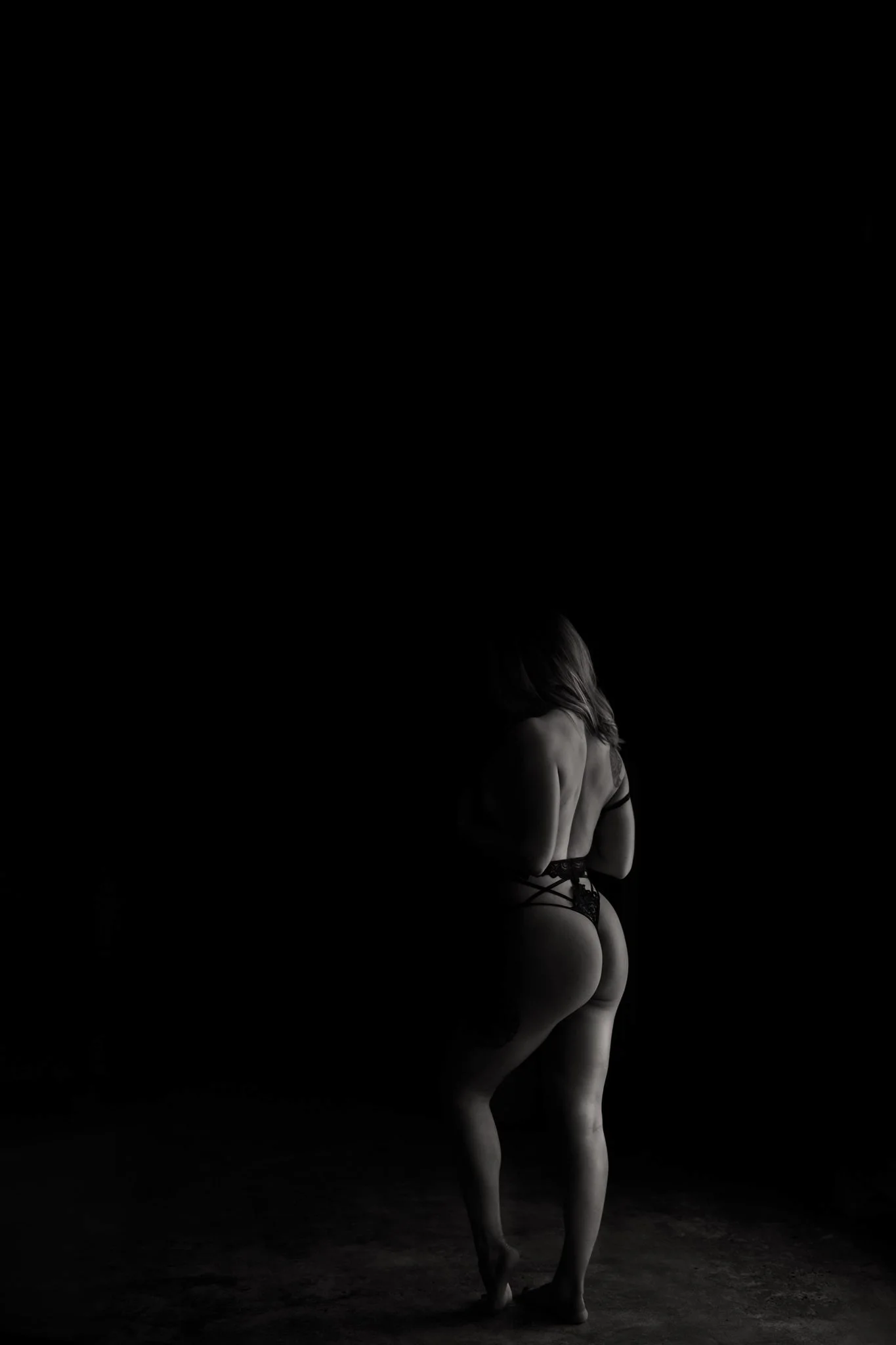 A woman with light skin and long hair, wearing black lace lingerie, posed against a dark background with minimal lighting highlighting her back and lower body.