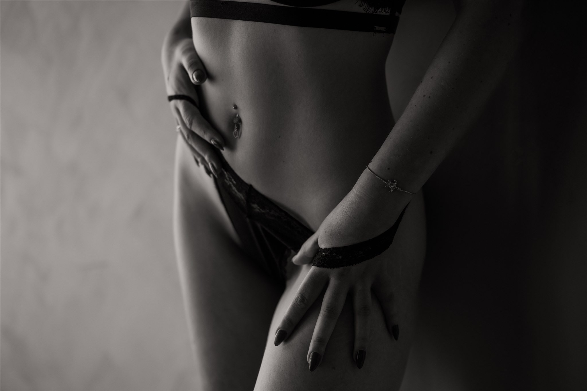A black and white photograph of a woman's midsection. She is wearing a crop top and underwear, with her hands positioned near her waist and thigh, showcasing jewelry including a belly button piercing, a bracelet, and rings. The lighting creates shado