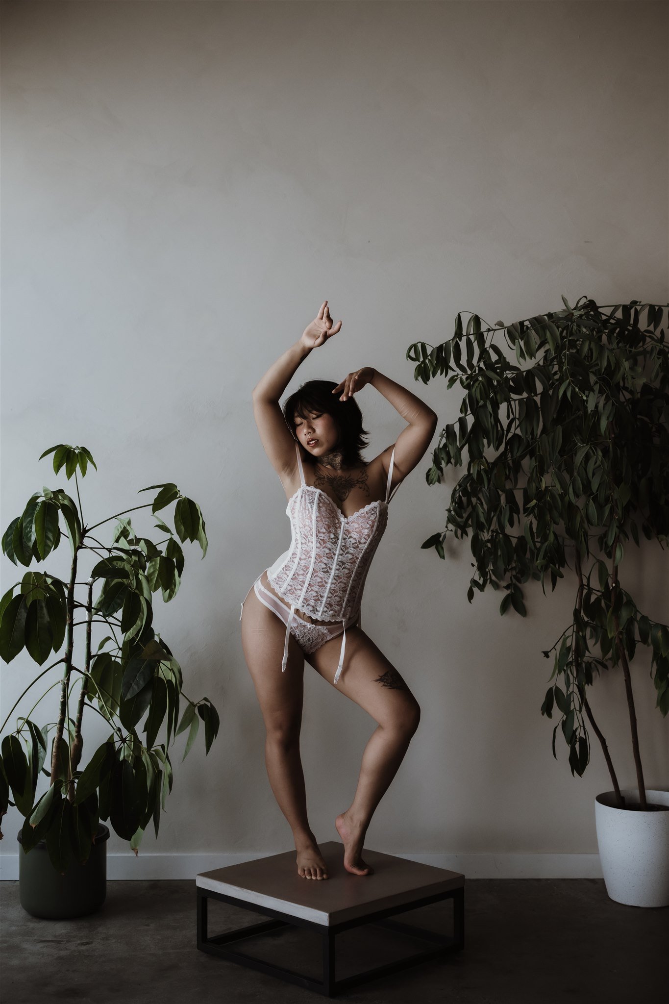 A woman in white lace lingerie dancing on a small platform in a minimalist room with two large potted plants.