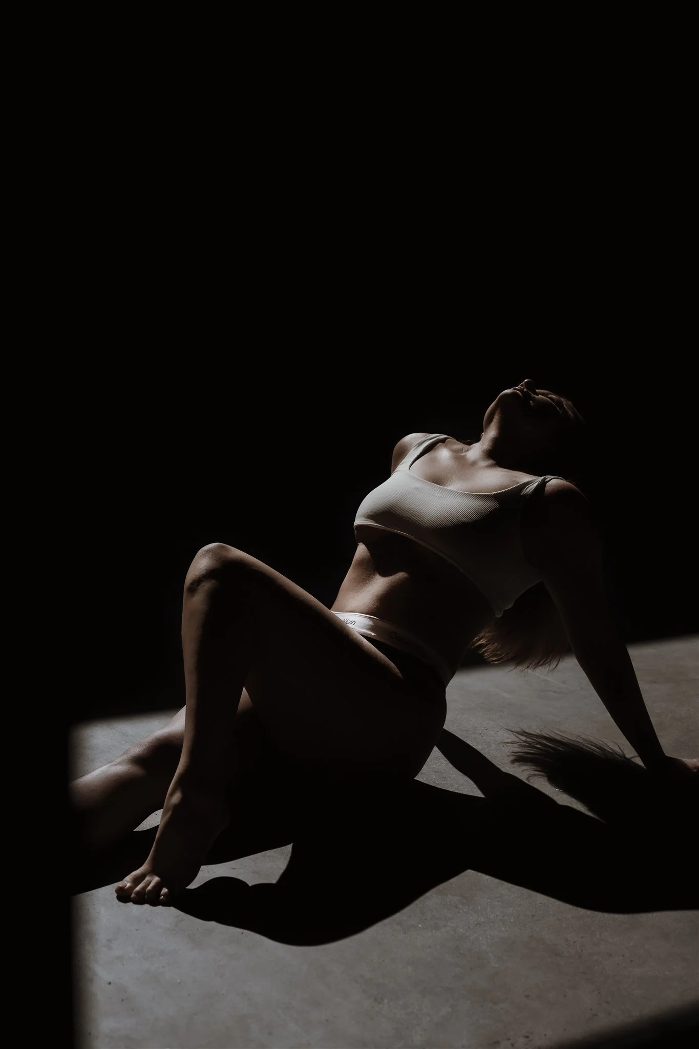 A woman in athletic wear posing on the floor with dramatic lighting creating shadows.