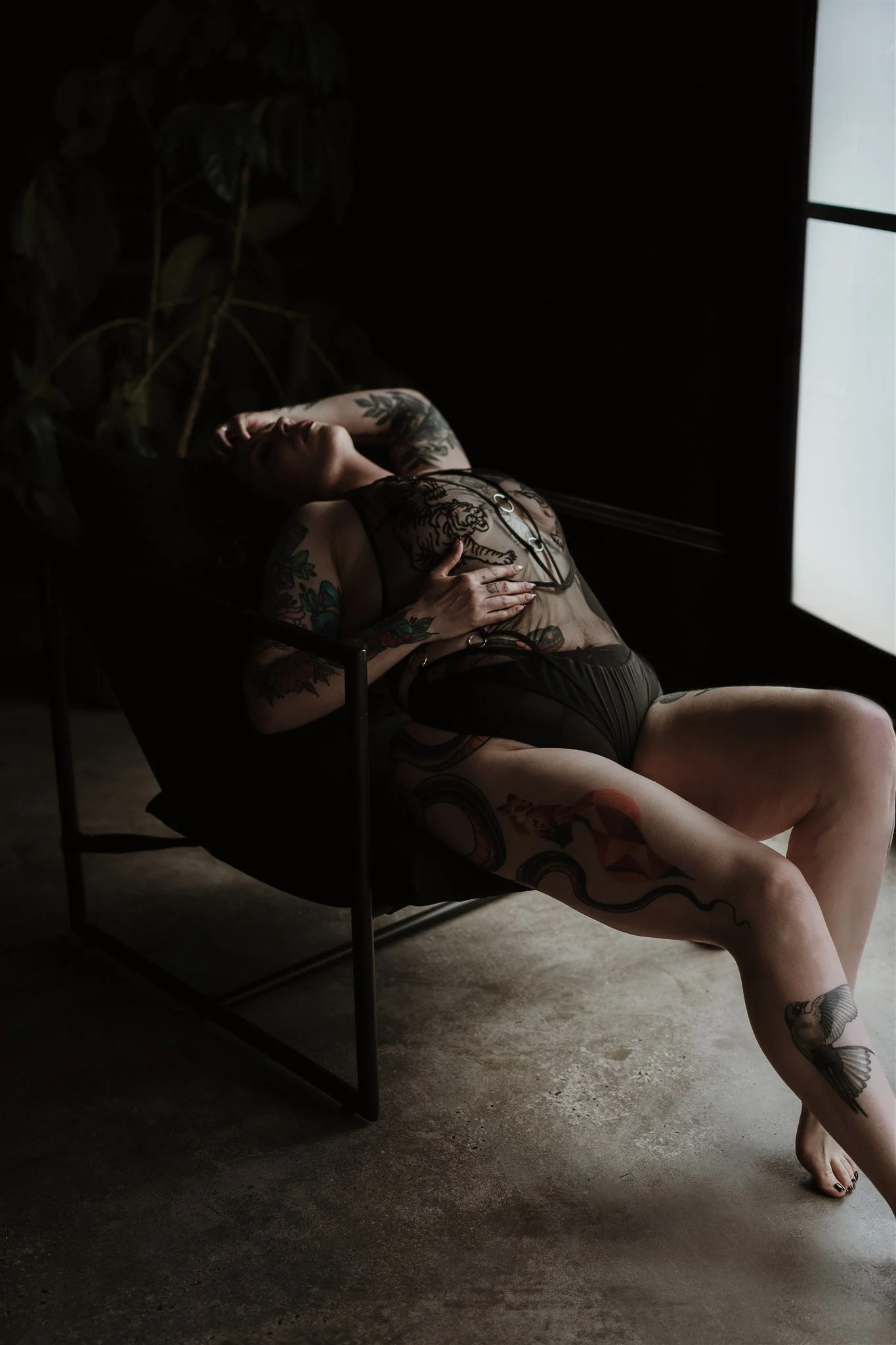 A tattooed woman in black lingerie reclining on a black chaise lounge, posed with her eyes closed and one hand behind her head, in a dimly lit room with a large window on the right.