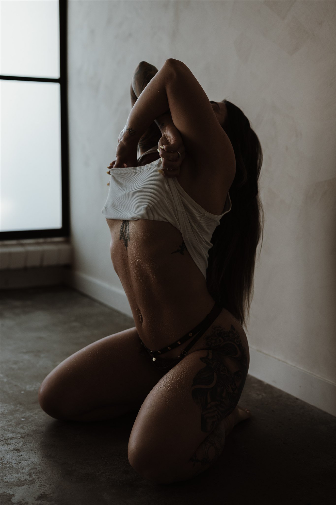 A woman with tattoos kneeling on the floor, lifting her shirt to reveal her midsection. She is in a room with a large window, with natural light illuminating her figure.