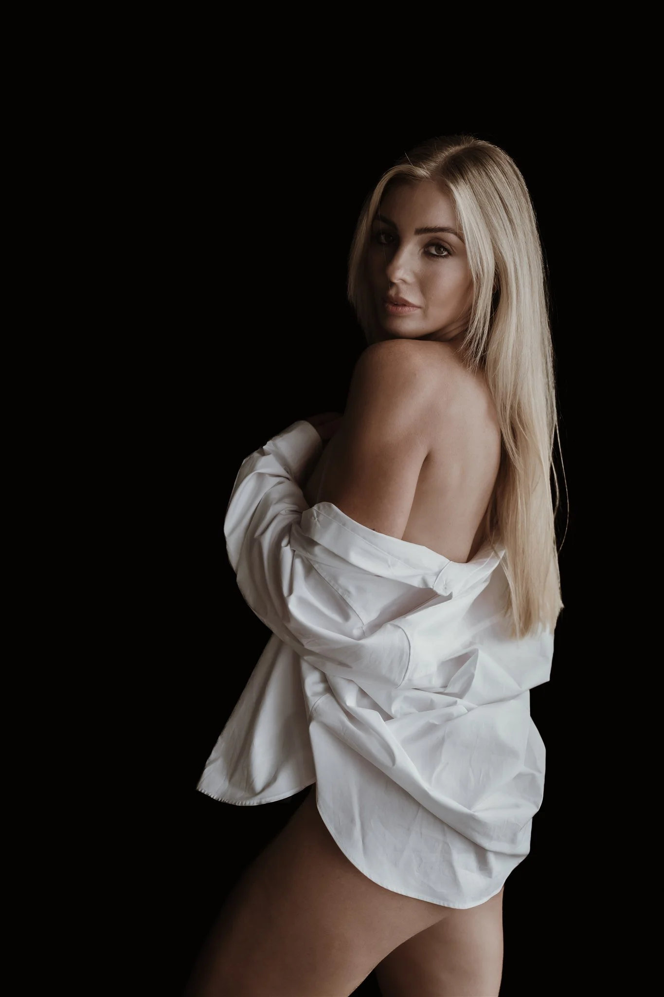 A woman with blonde hair, looking over her shoulder, partially draped in a white shirt, against a black background.