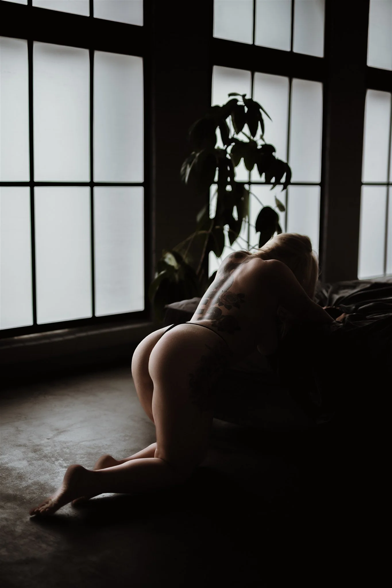 A woman with tattoos is kneeling on the floor and resting her head on a bed in front of large windows, with a plant in the background.