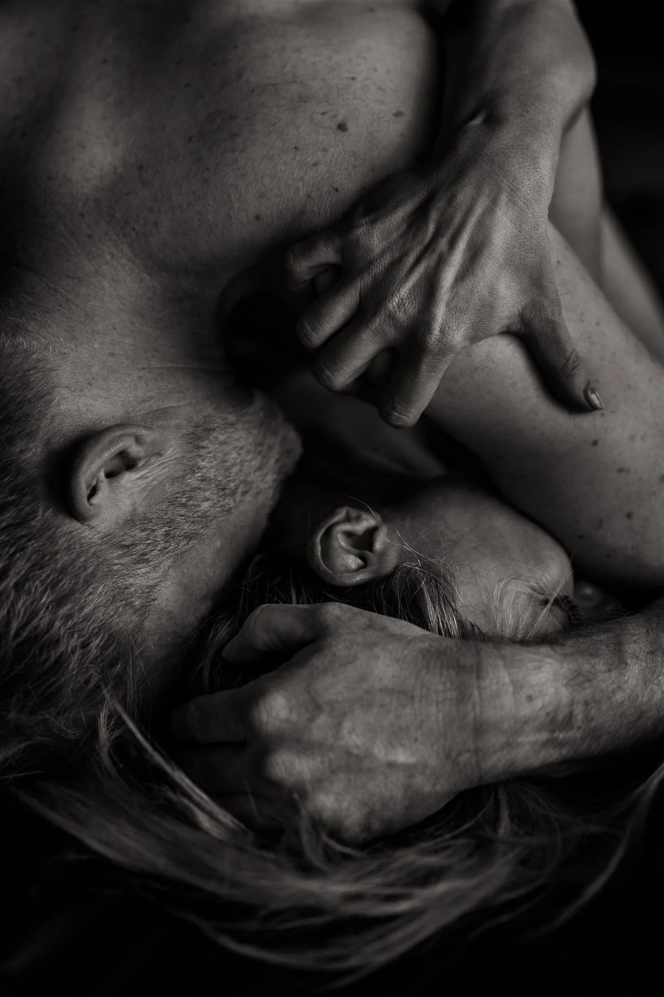Black and white close-up of two people embracing, with one person's head resting on the other's chest, and their hands gently holding each other's heads, conveying intimacy and tenderness.