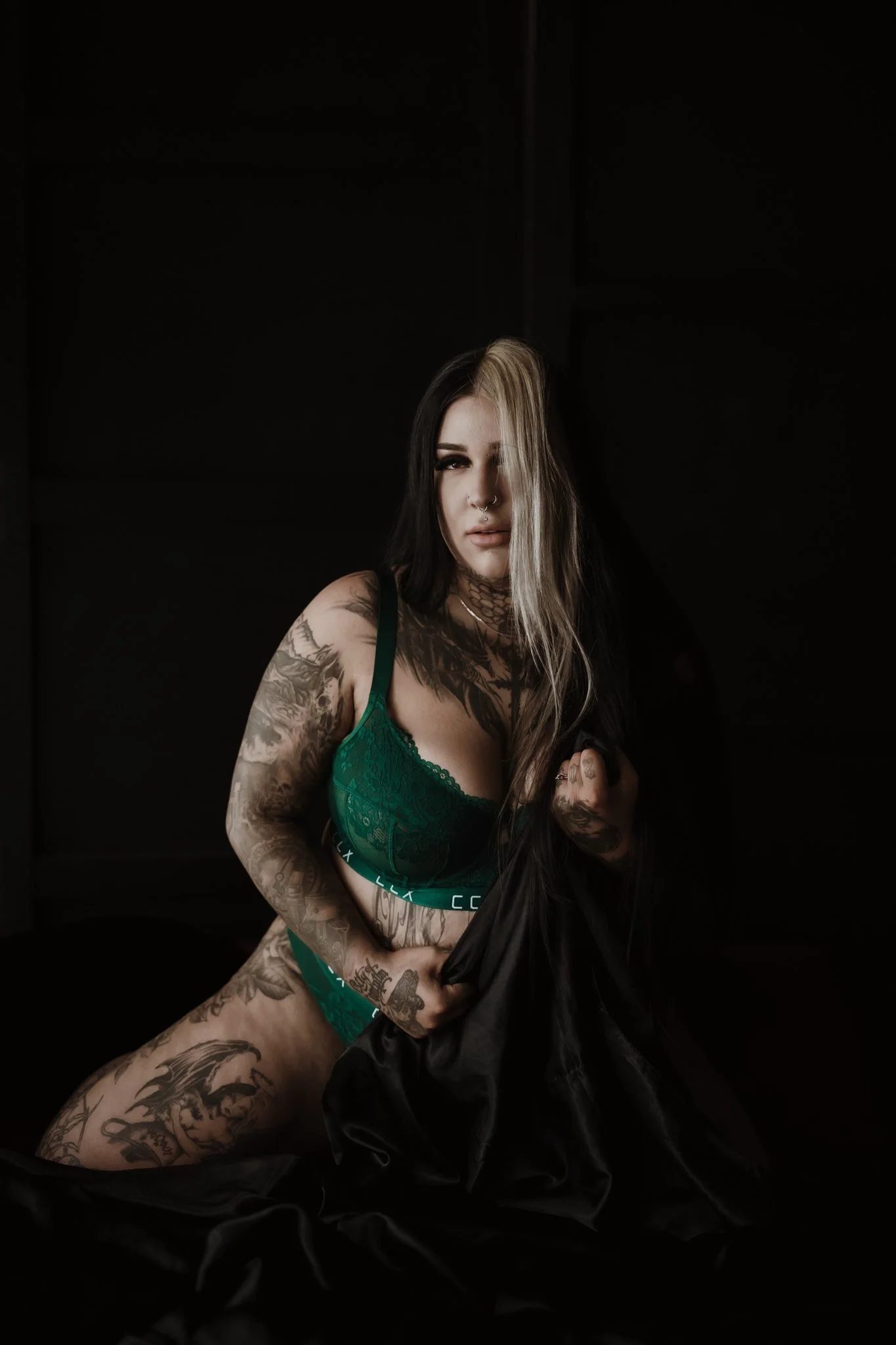 A tattooed woman with long black and blonde hair wearing a green lace bra poses in a dark room, holding a black fabric.