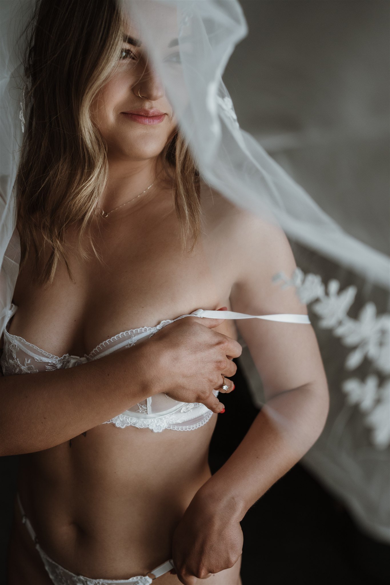 A woman with shoulder-length wavy hair, wearing a sheer veil and white lace lingerie, is covering her chest with her hand, and looking softly to the side.