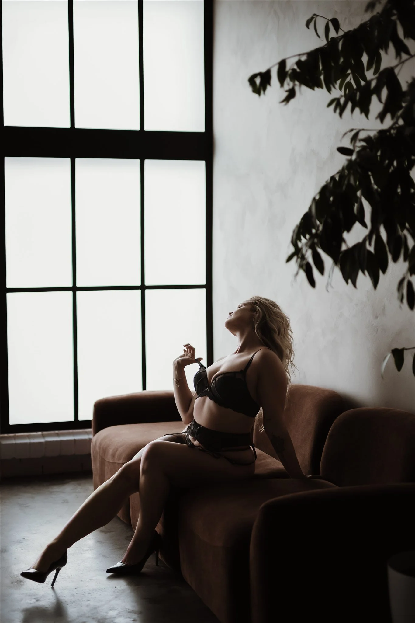 A woman with wavy blonde hair wearing black lingerie and high heels sitting on a brown sofa in front of a large window with black framing, with a white wall and a plant nearby.