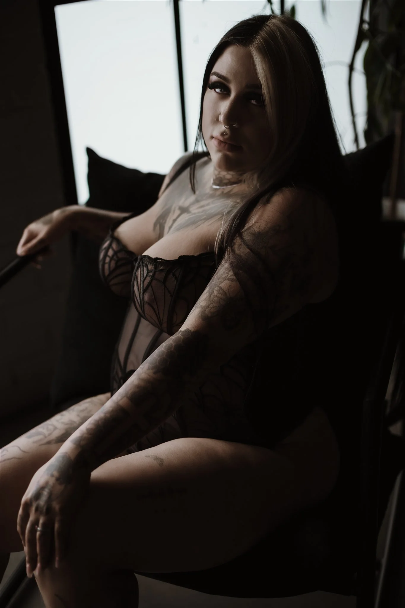 A woman with dark hair and tattoos wearing black lacy lingerie sitting on a chair in a dimly lit room, with a bright window in the background.