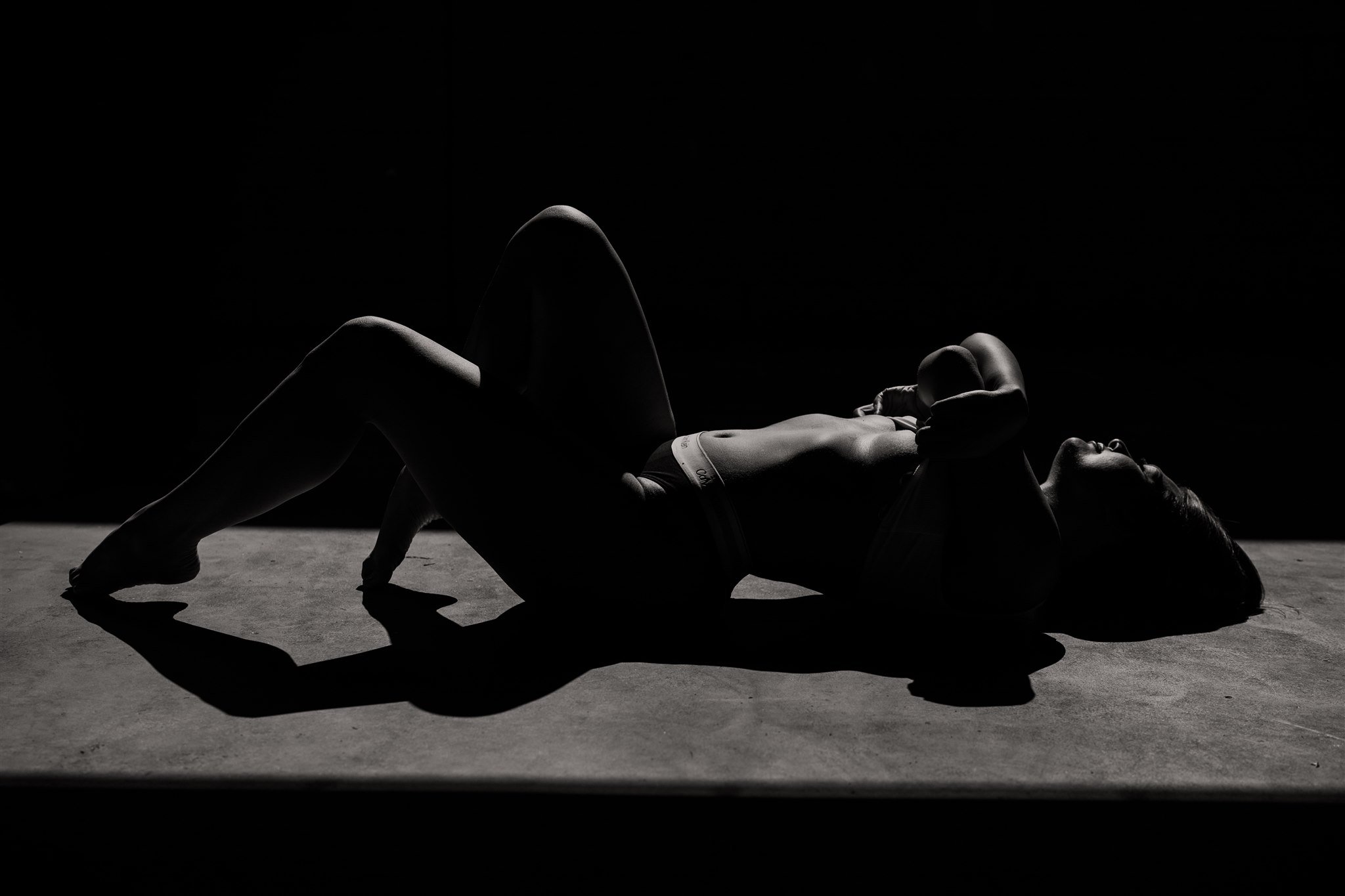 A silhouette of a woman lying on the floor with one leg bent and her hands behind her head, in a dimly lit setting.