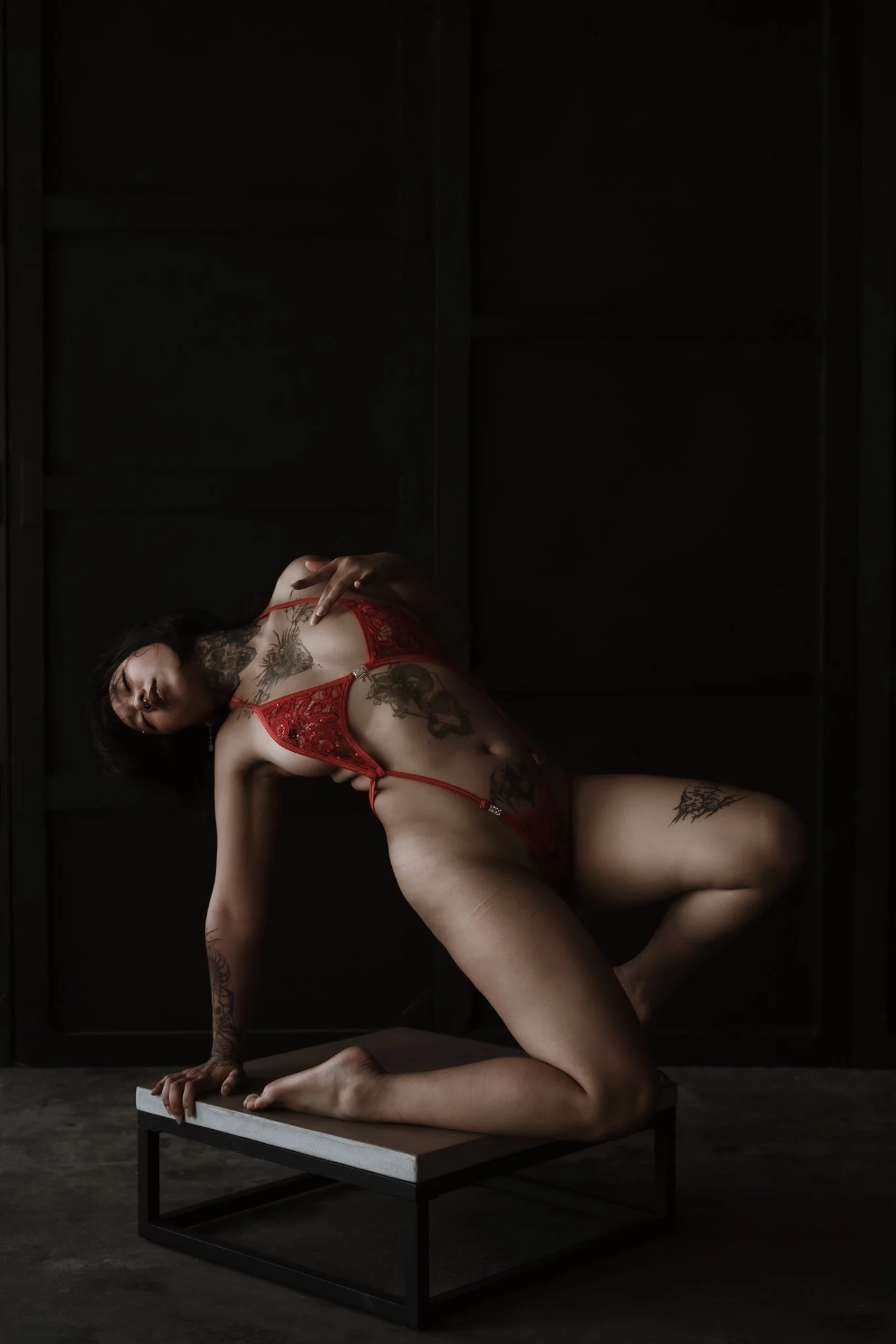 A woman with tattoos wearing red lingerie, posing on a small table in a dark room.