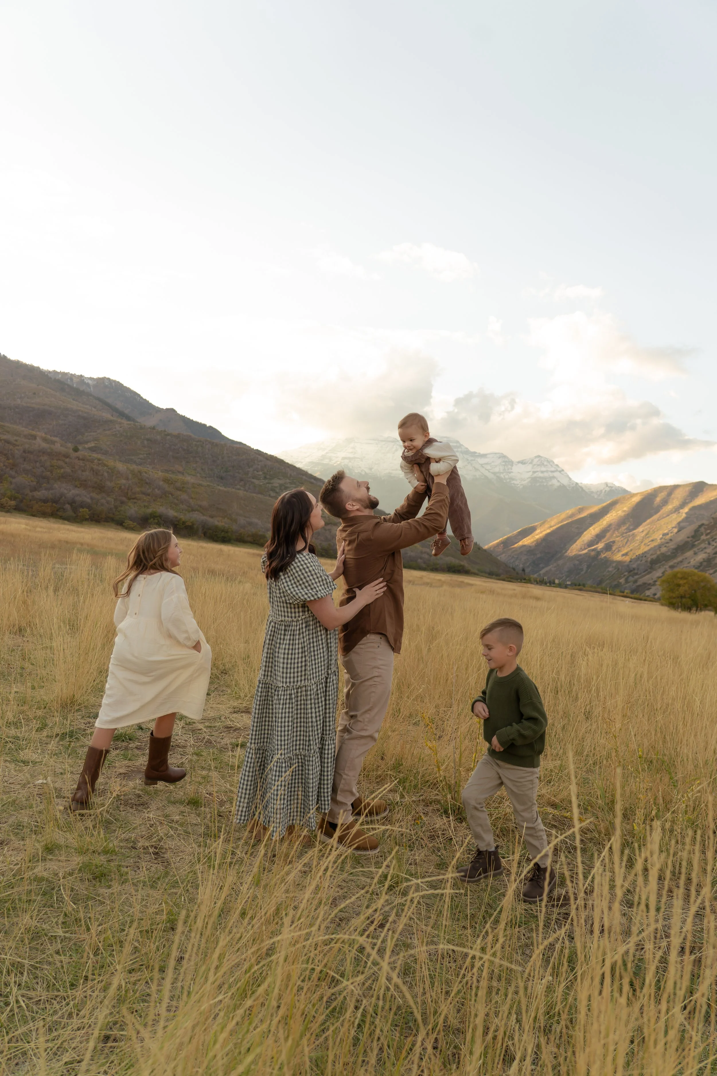 A family of five enjoying a day outdoors in a grassy field with mountains in the background. The father is lifting a young boy into the air, while the mother and two other children watch and smile.