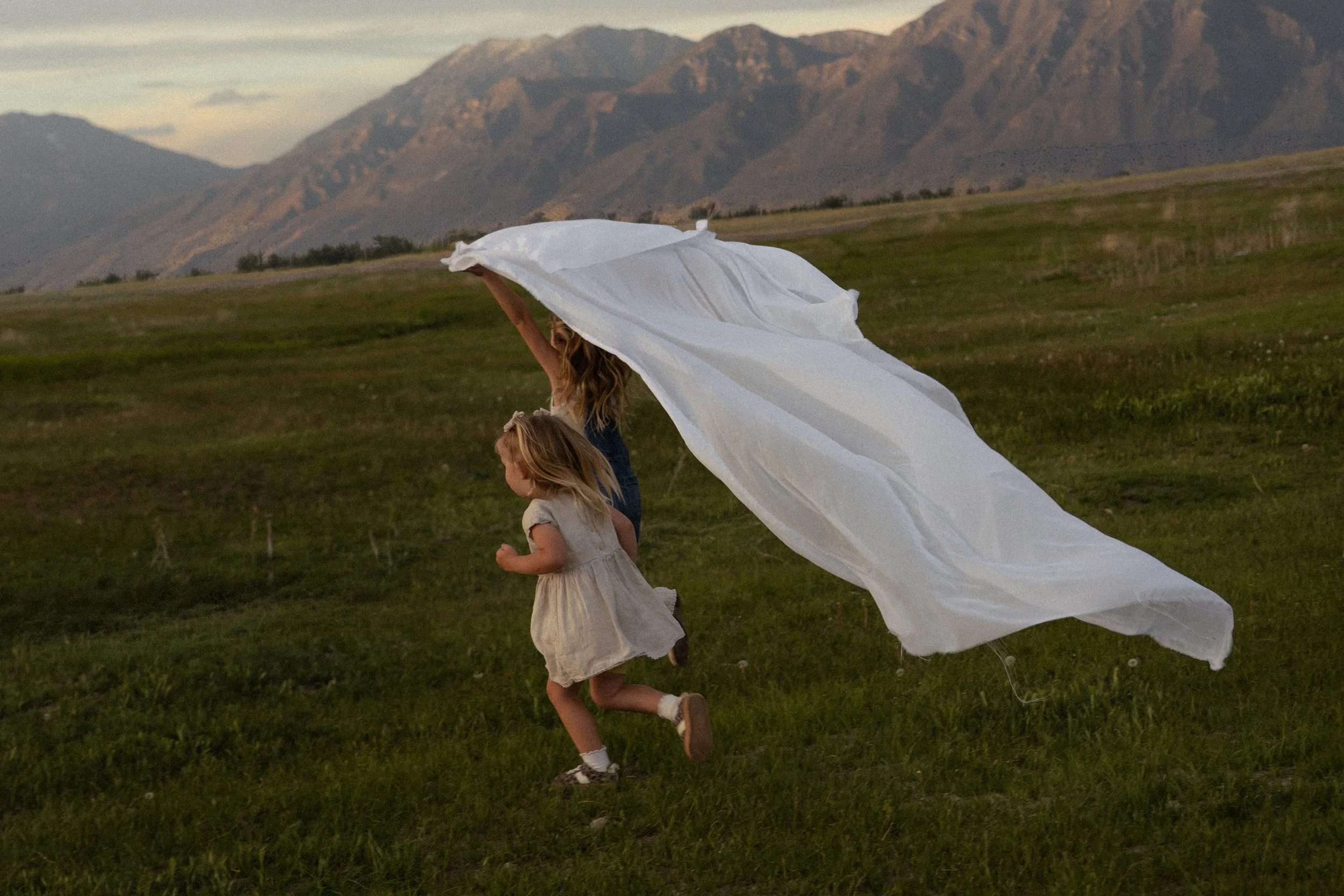 A woman and two young girls run across a grassy field with mountains in the background. The woman holds a flowing white sheet or fabric above her head, creating a sense of movement.