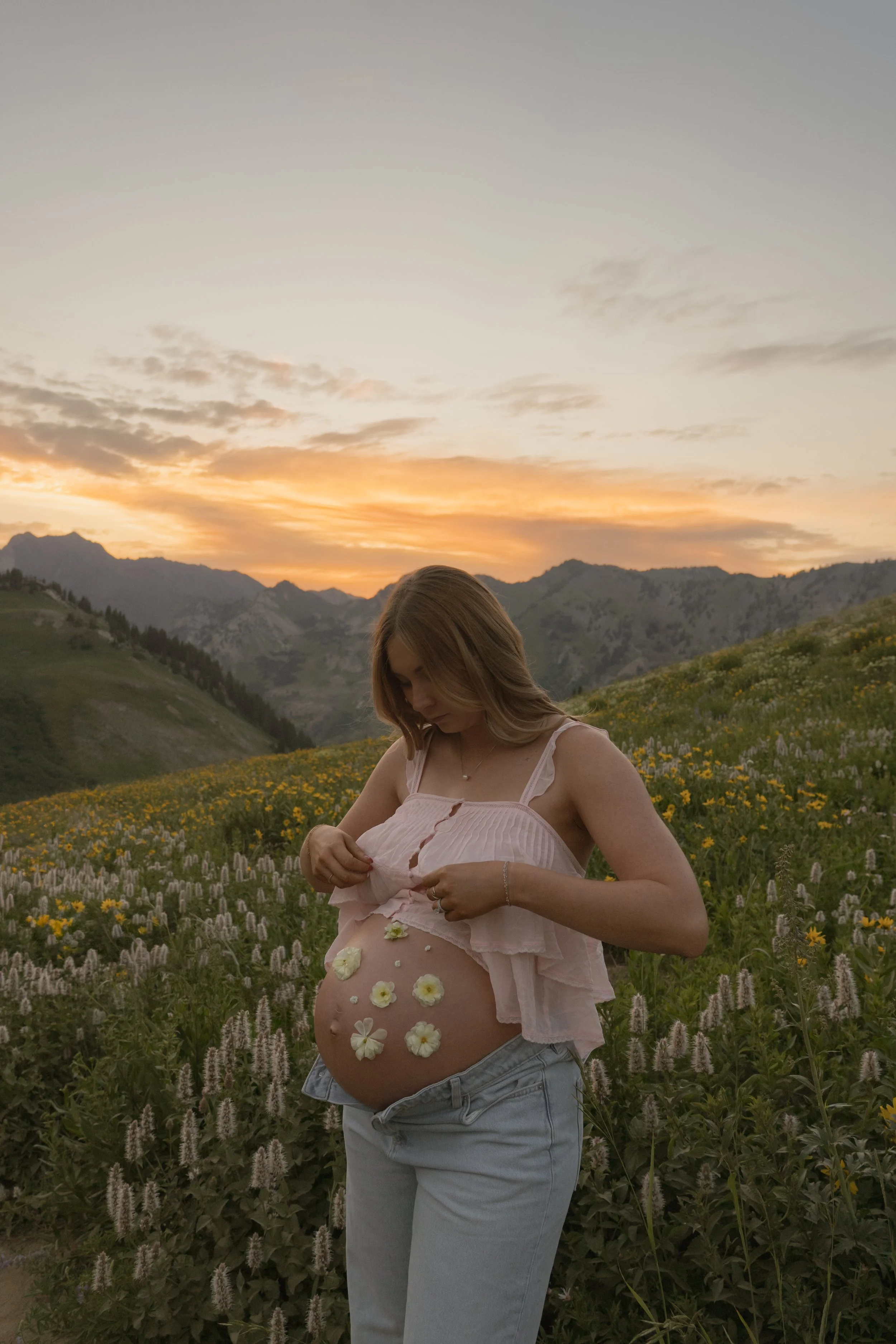 Pregnant woman standing in a field of flowers at sunset, wearing a light pink top and jeans, with flowers on her belly.