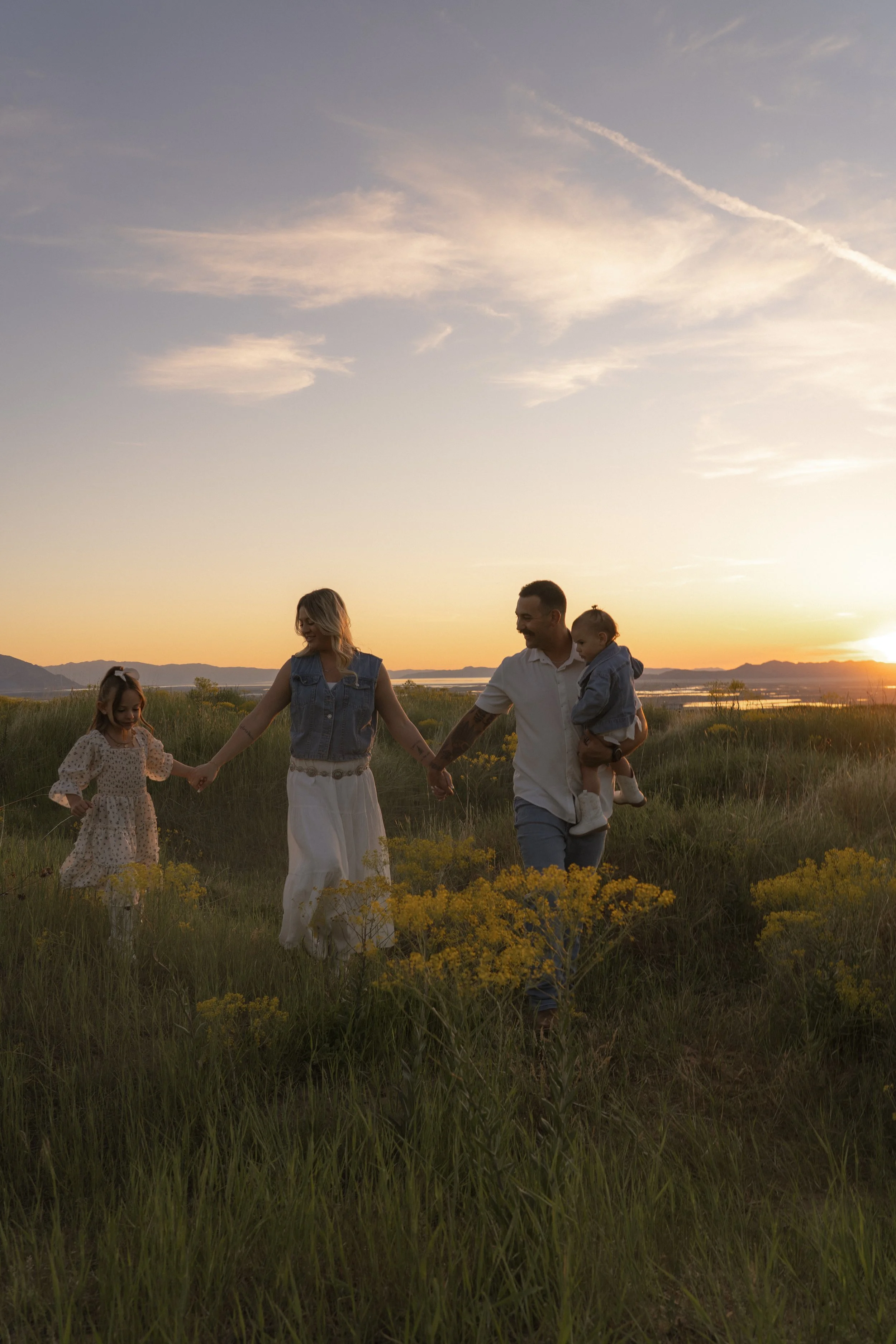 A family of four holding hands and walking through a grassy field during sunset, with mountains and a body of water in the background.