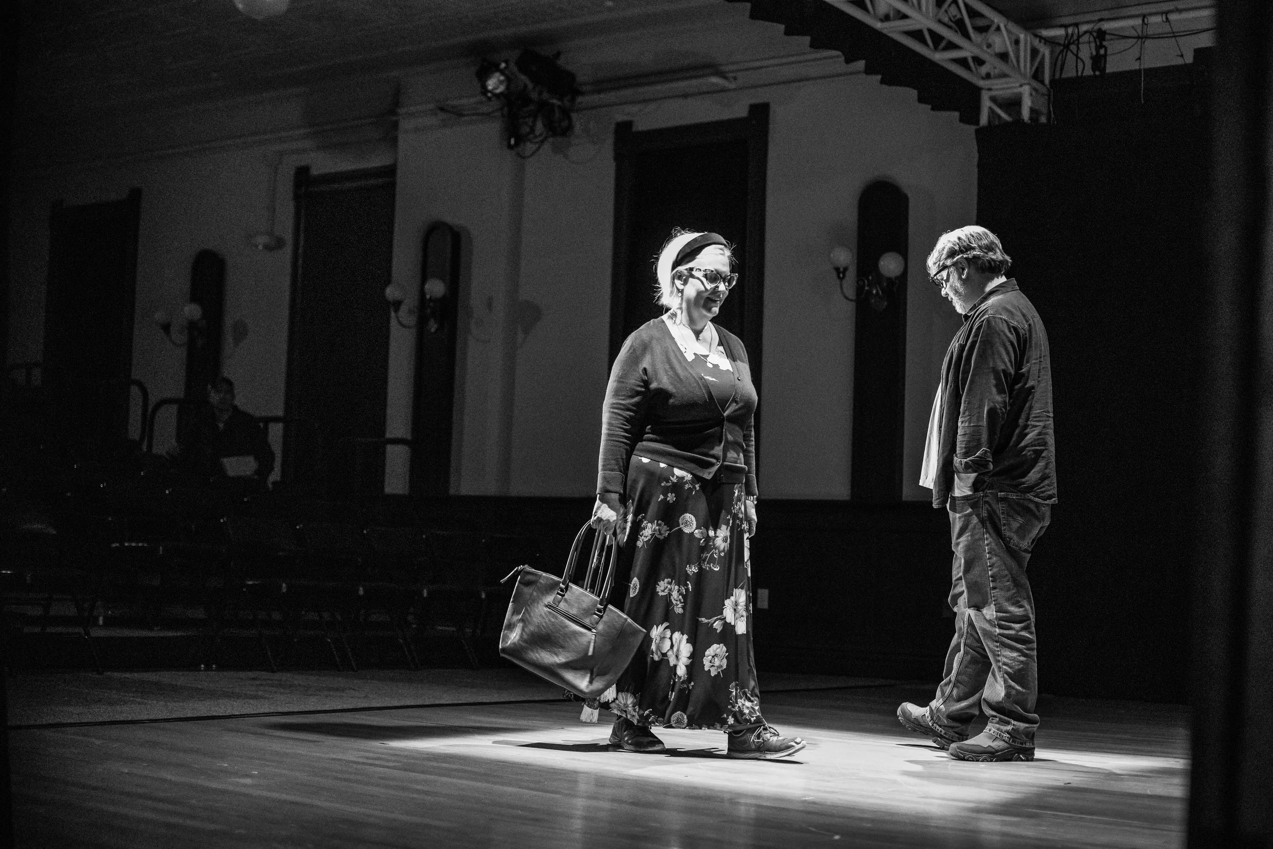 Two people standing on a stage in a theater, engaged in a conversation. The woman is wearing a floral skirt, glasses, and holding a large handbag. The man is looking down, wearing a jacket and jeans.