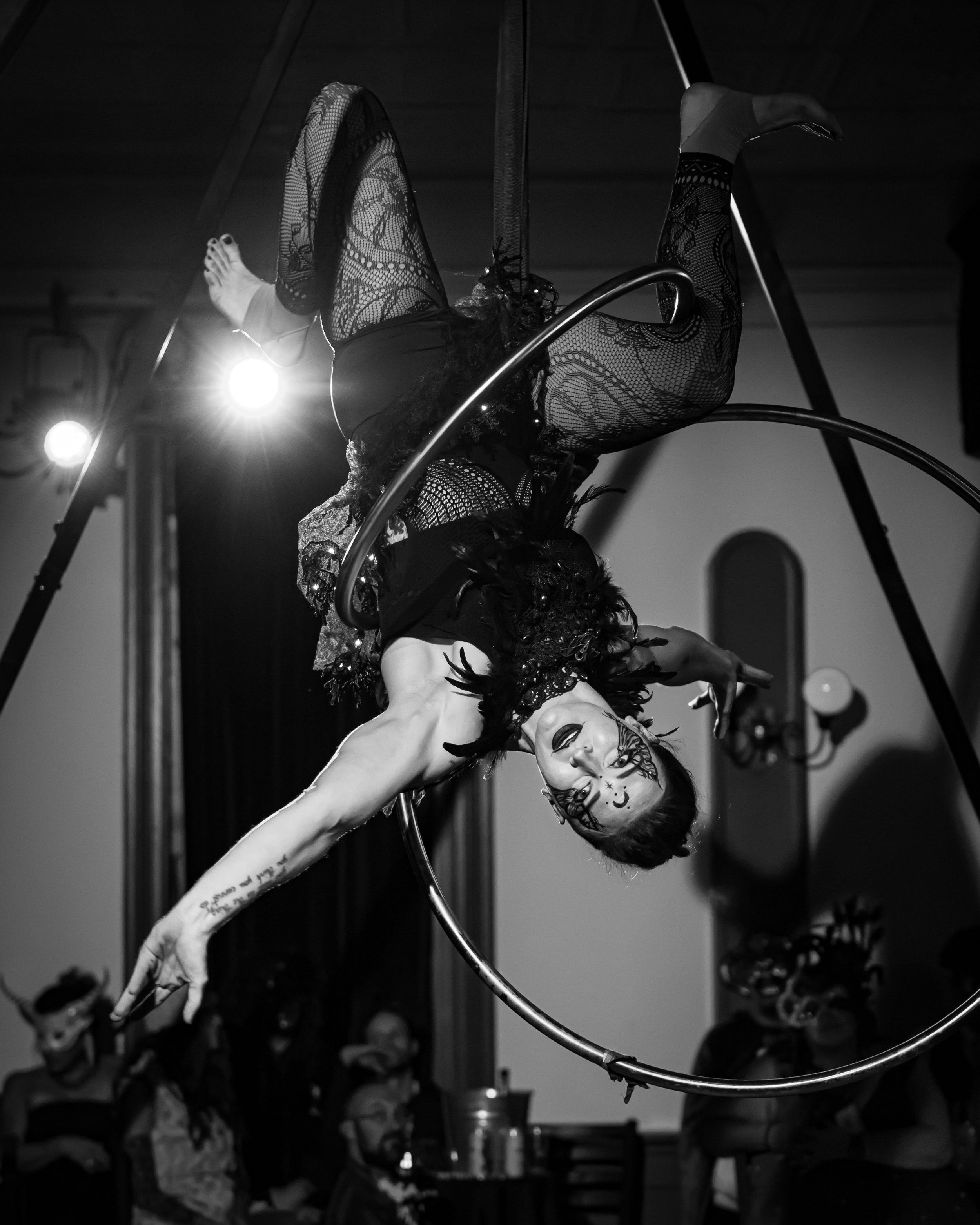 An aerial performer in intricate costume, hanging upside down on a hoop, with face makeup, in a performance setting.