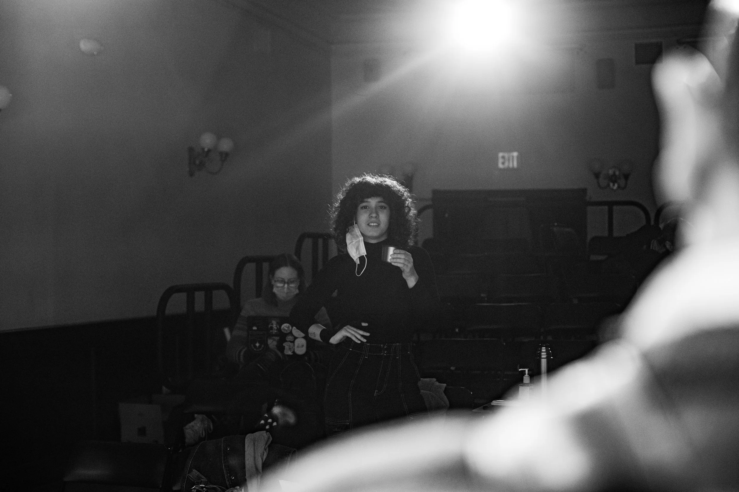 A woman with curly hair holding a cup standing in a dimly lit room, with another woman seated in the background wearing a face mask and looking at a laptop.