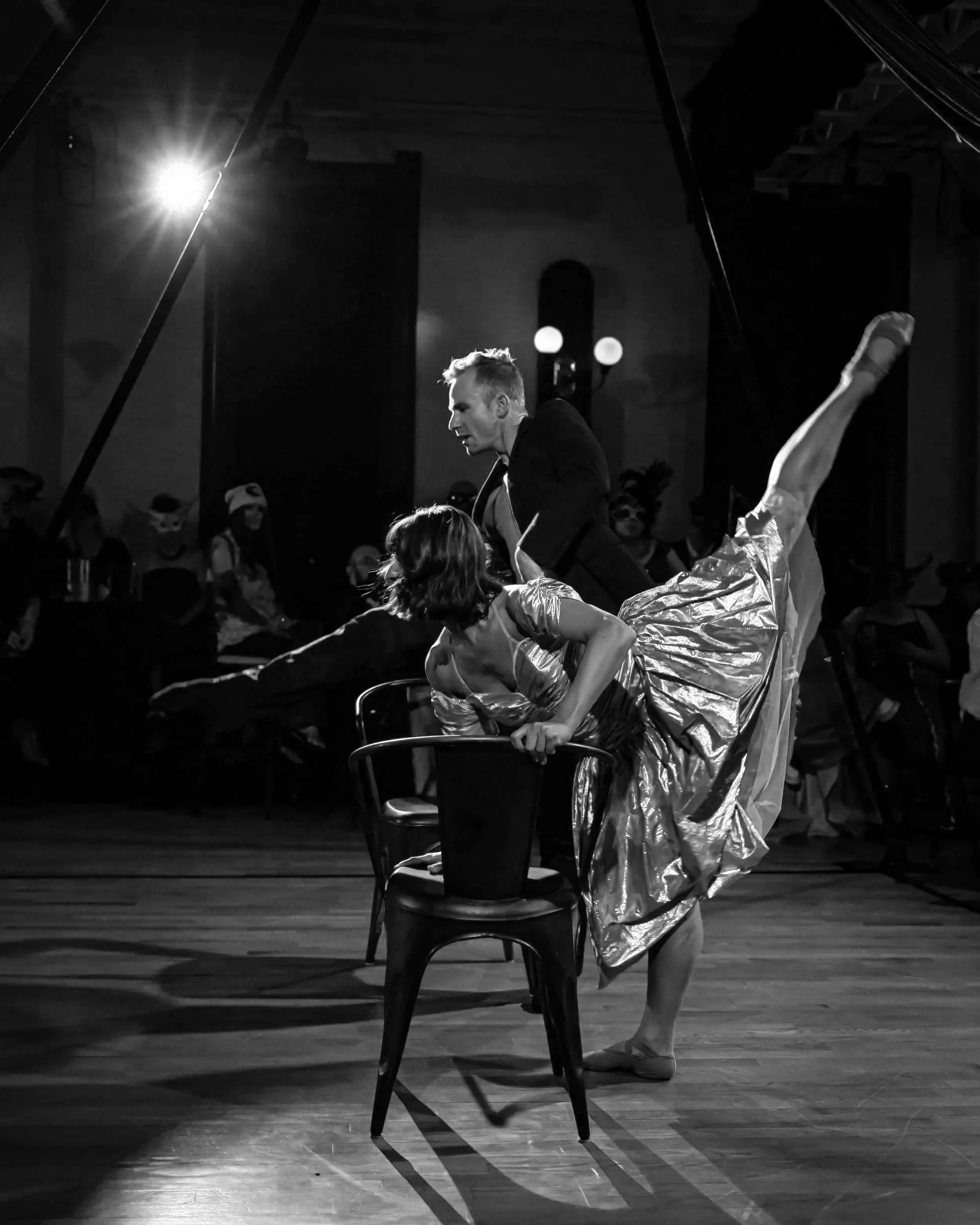 A black and white photo of two performers, one man and one woman, dancing and posing on a stage with an audience in the background. The woman is wearing a shiny dress and balancing on one leg with her other leg extended high, while leaning on a chair.