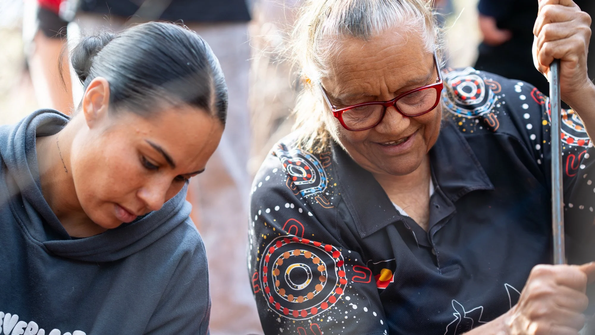 The Elder and the woman in the hoodie are both staring down at the ground. The Elder smiles, both of her hands holding onto the pole.