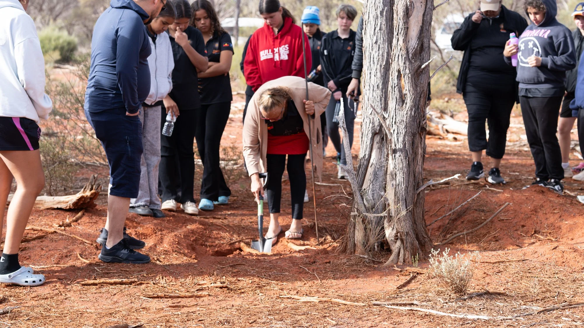 The Elder bends down near a grey tree in the middle of the photograph and digs with a short shovel into the red dirt. Her other hand holds onto a thin pole The teenagers watch from around her.