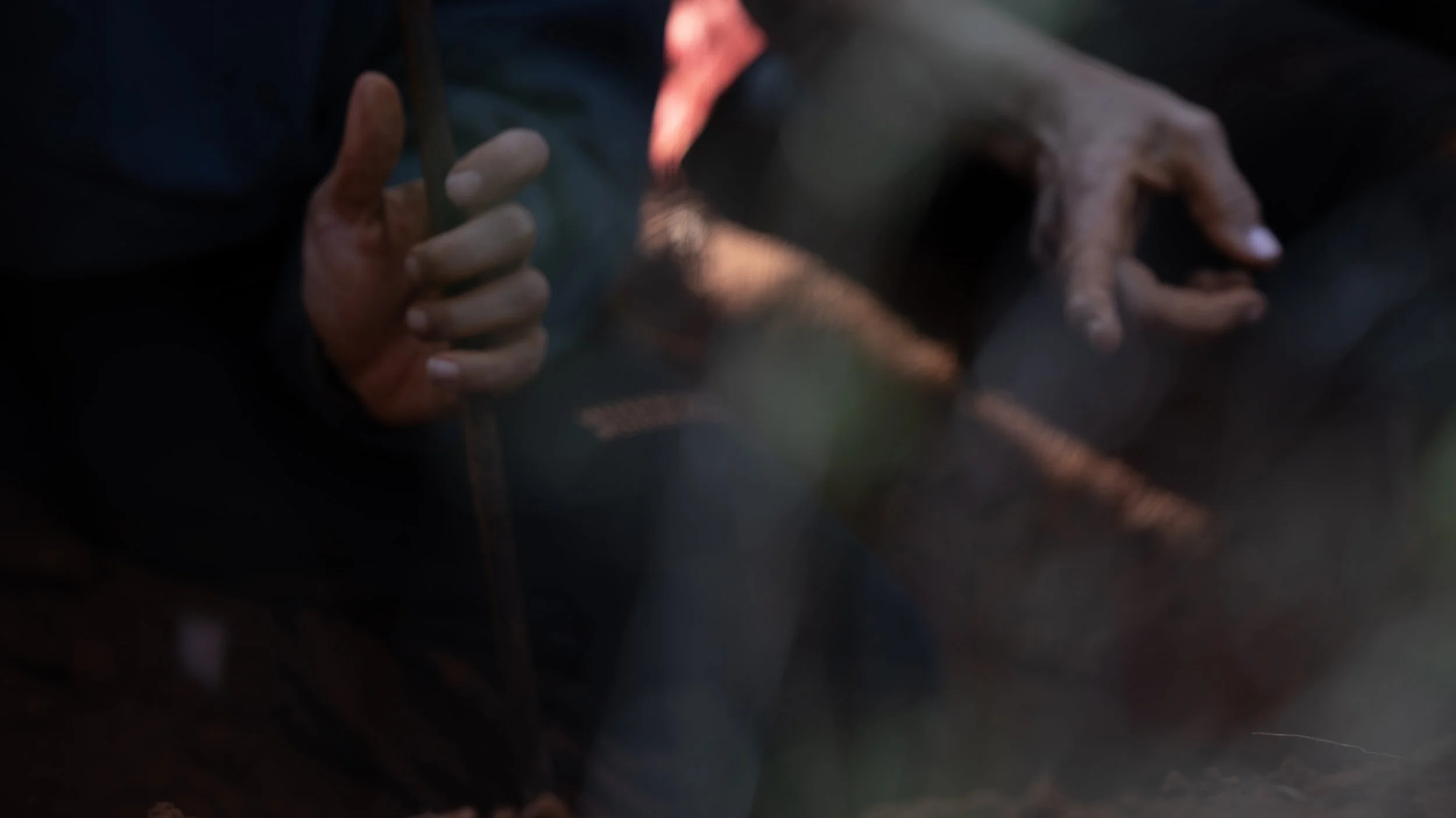 The woman in the hoodie holds onto the pole on the left-hand side of the frame as the Elder points at the dirt on the right-hand side of the frame. The image is slightly out of focus.