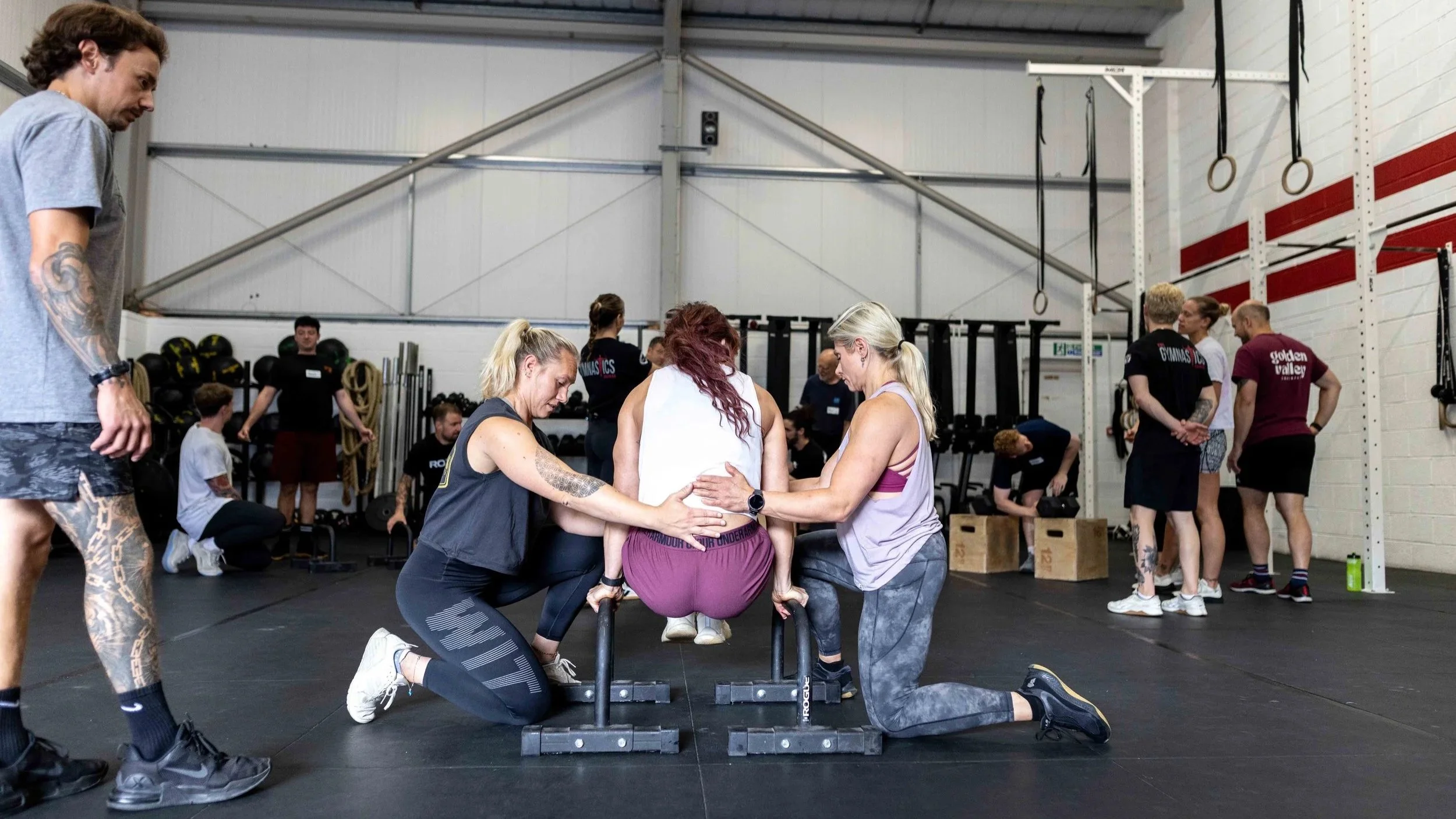 A woman is performing a handstand on parallel bars in a gym, with two women supporting her by holding her waist and legs.