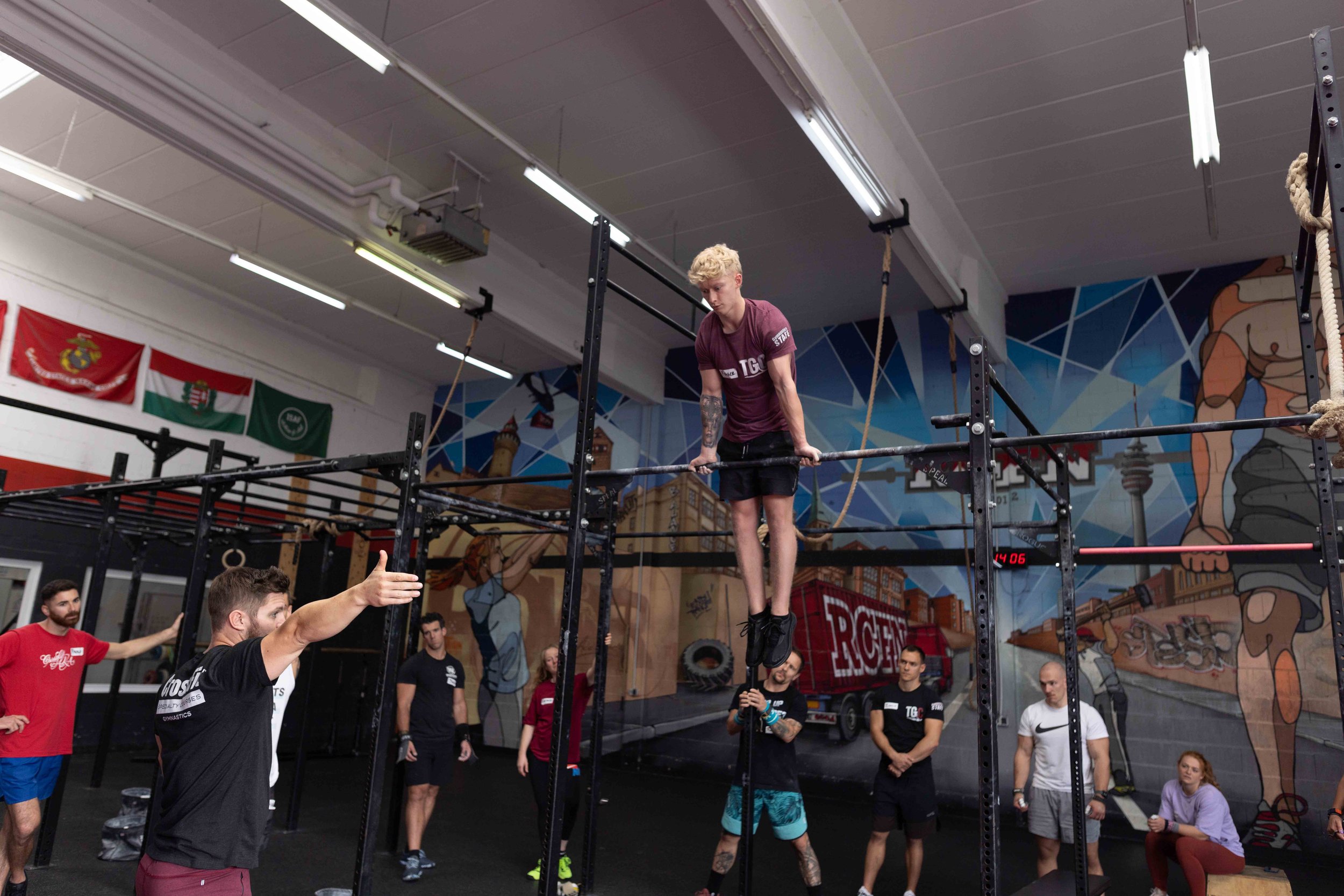 A man performing a gymnastic move on a bar during a training session at a gym with a colorful mural on the wall.
