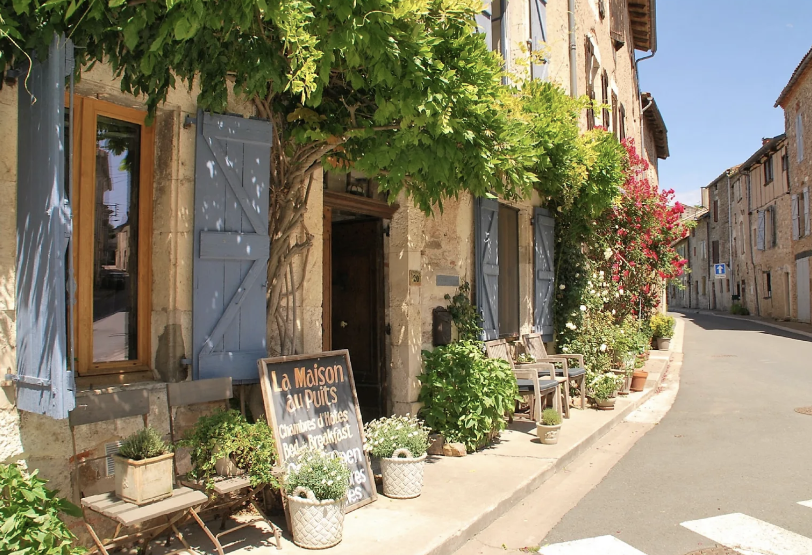 A charming French street scene with a stone house featuring blue shutters, potted plants, a seating area, and a colorful flowering shrub. A chalkboard sign advertises rooms and breakfast, and the street curves to the right with more stone buildings and an upward slope.