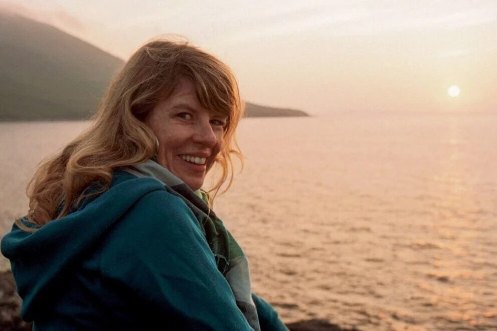 Woman with red hair smiling at sunset over a body of water with hills in the background.