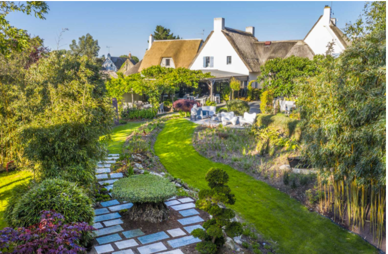Unique delicious restaurant recommendations in France - A lush, landscaped garden with a curved grass pathway, surrounded by various plants and flowers, in front of a house with a thatched roof and white exterior, under a clear blue sky.