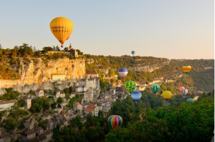 Discover Rocamadour - Multiple colorful hot air balloons floating over a hillside village with trees and buildings in the background.