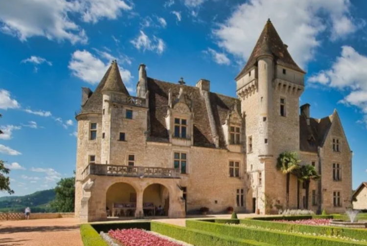 Josephine Baker's Castle in the Dordogne region - a historic stone castle with four round towers, multiple windows, and ornate details, set against a blue sky with scattered clouds.