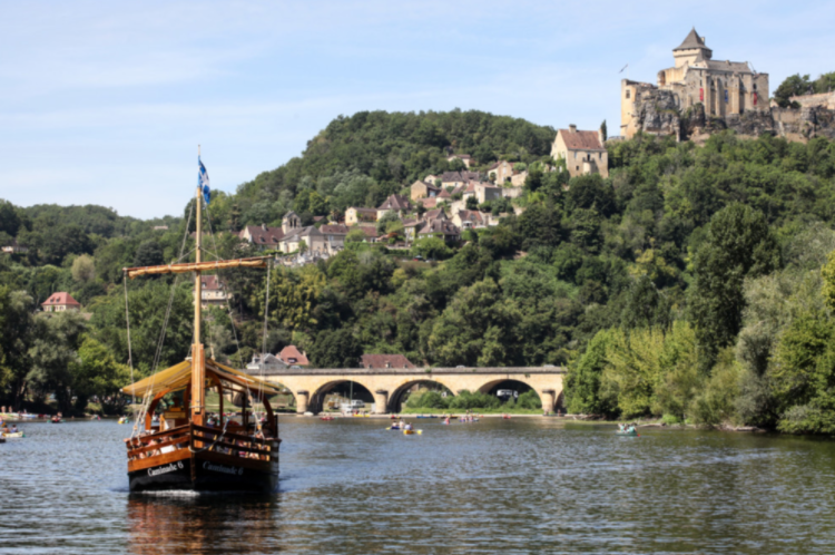 Dordogne river adventures - a wooden sailboat and several small boats, surrounded by lush green trees and a hillside with houses, a stone bridge, and a castle on top.
