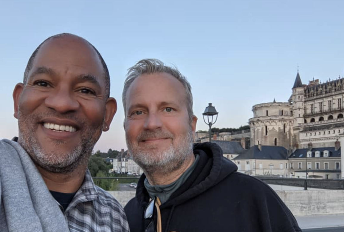 Two men smiling for a photo outdoors with a historic castle or palace in the background during sunset.