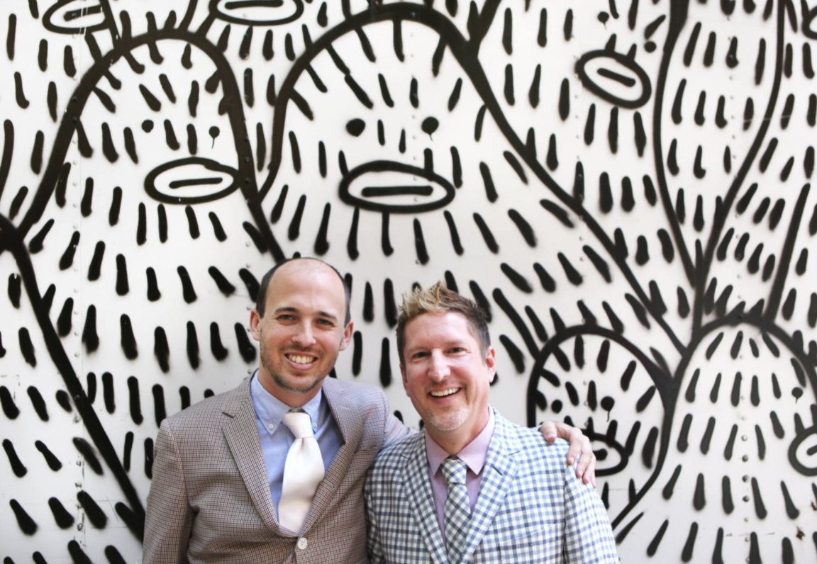 Two men smiling and standing together in front of a black and white abstract mural with bold, thick lines and stylized leaf shapes.